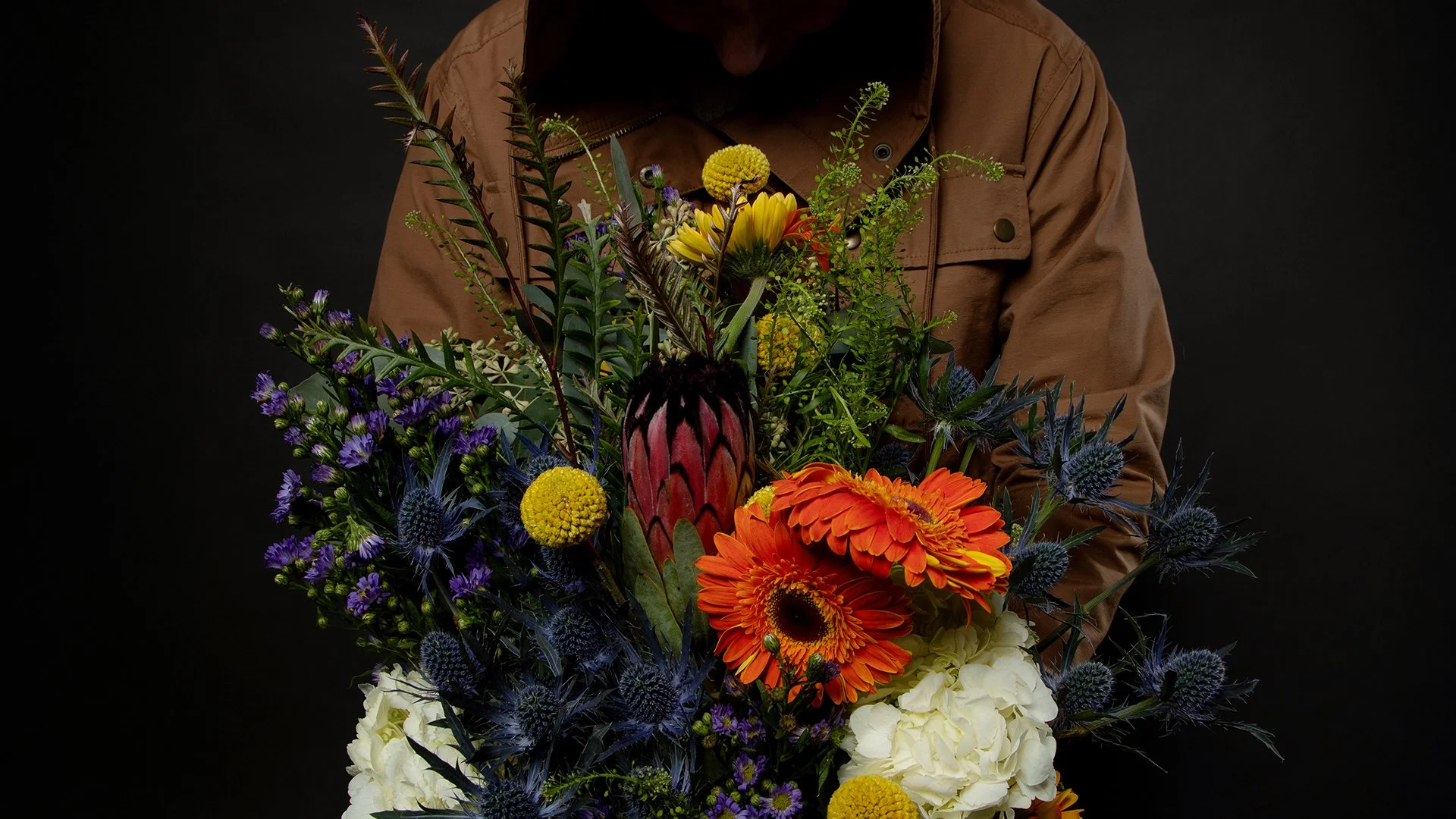 Person holding a colorful bouquet of flowers against a dark background, with visible brown jacket and face partially obscured.