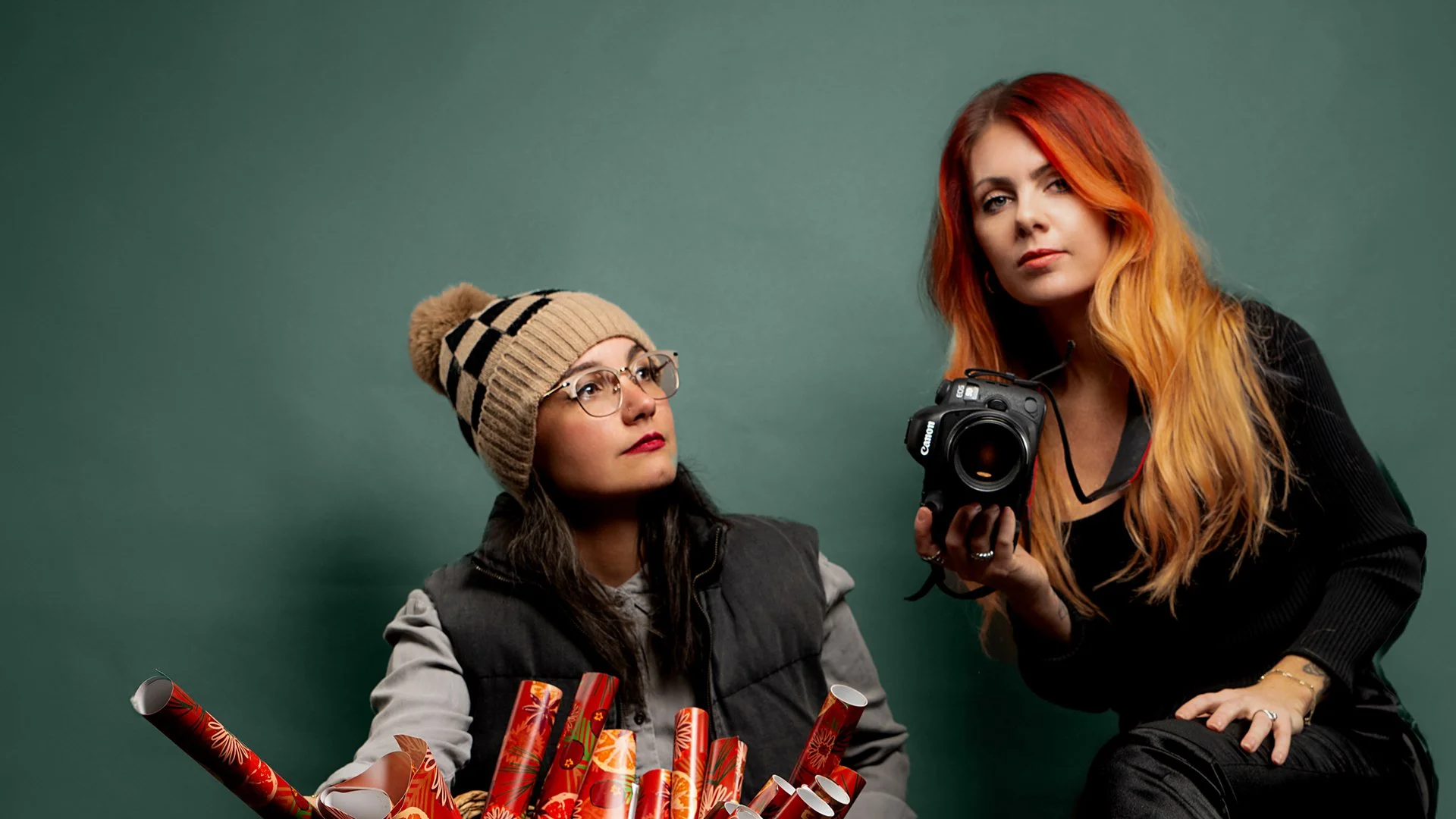 Two women, one wearing glasses and a striped beanie, sit with a basket of colorful party poppers. The other woman with long red hair holds a professional camera, wearing a black jacket, against a plain green background.