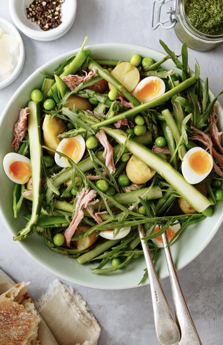 Green vegetable spring summer salad with halved soft-boiled quail's eggs, asparagus, potatoes, peas, and shredded ham hock in a white bowl.