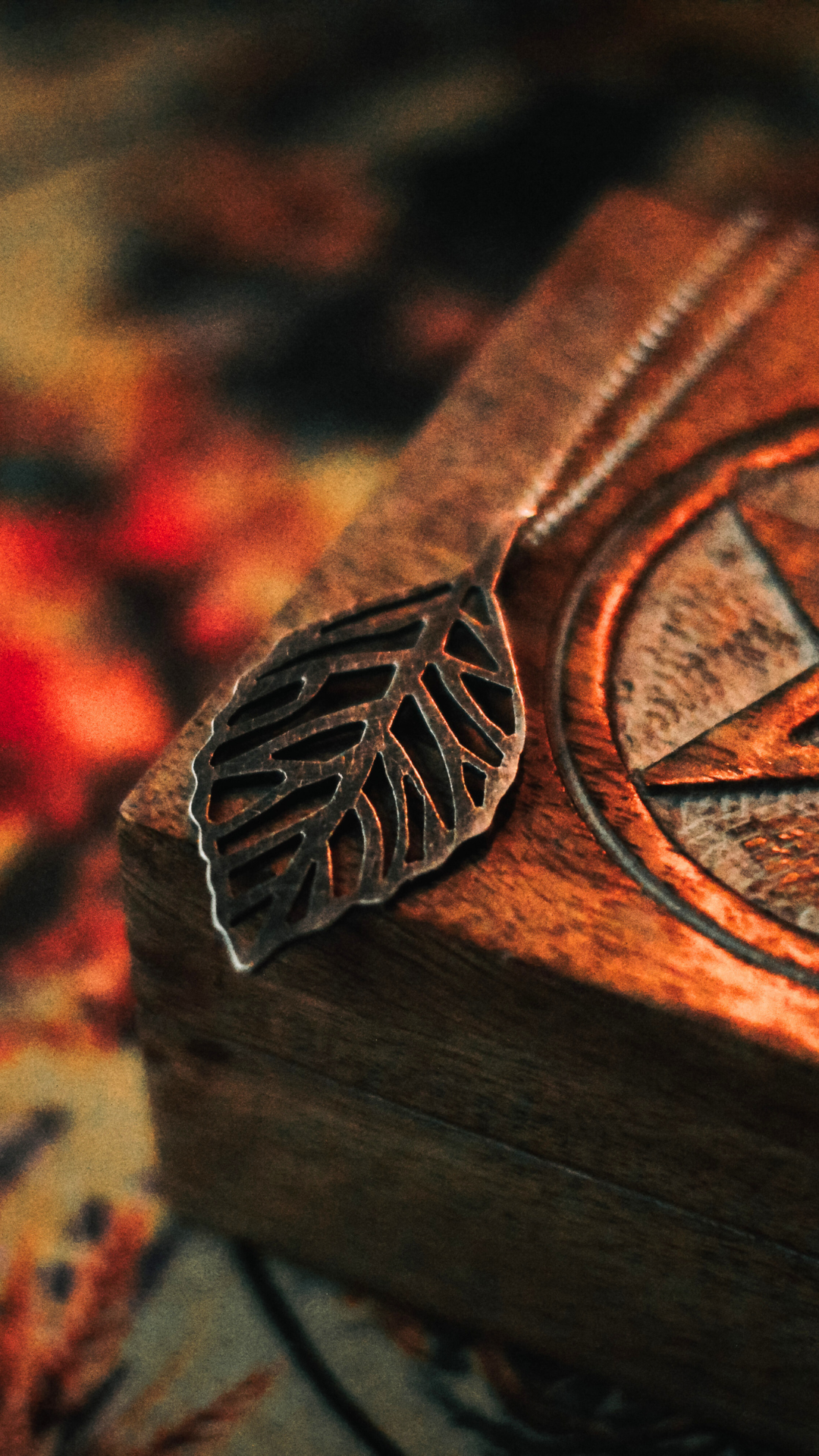 Close-up of a silver leaf-shaped jewelry charm resting on a wooden surface with a circular carved design. Blurred autumn leaves are visible in the background.