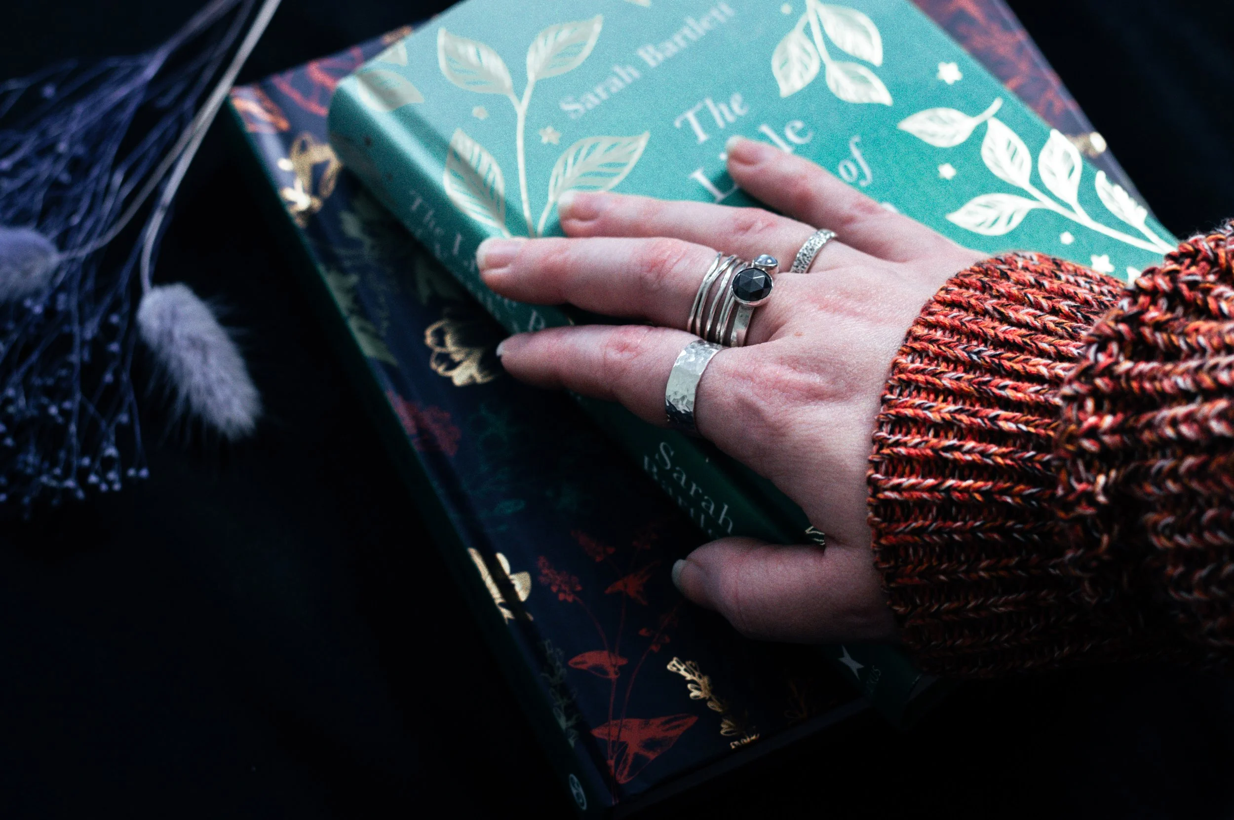 A close-up of a hand with multiple rings resting on a book titled 'The Solar System'. The person is wearing a multicolored knitted sweater, and there are decorative feathers nearby.