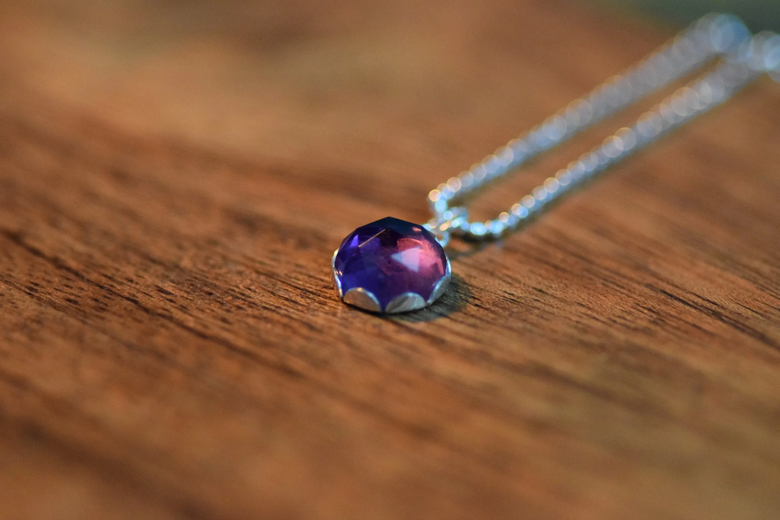 A close-up photograph of a purple, faceted gemstone pendant necklace resting on a wooden surface, with the chain extending into the background.