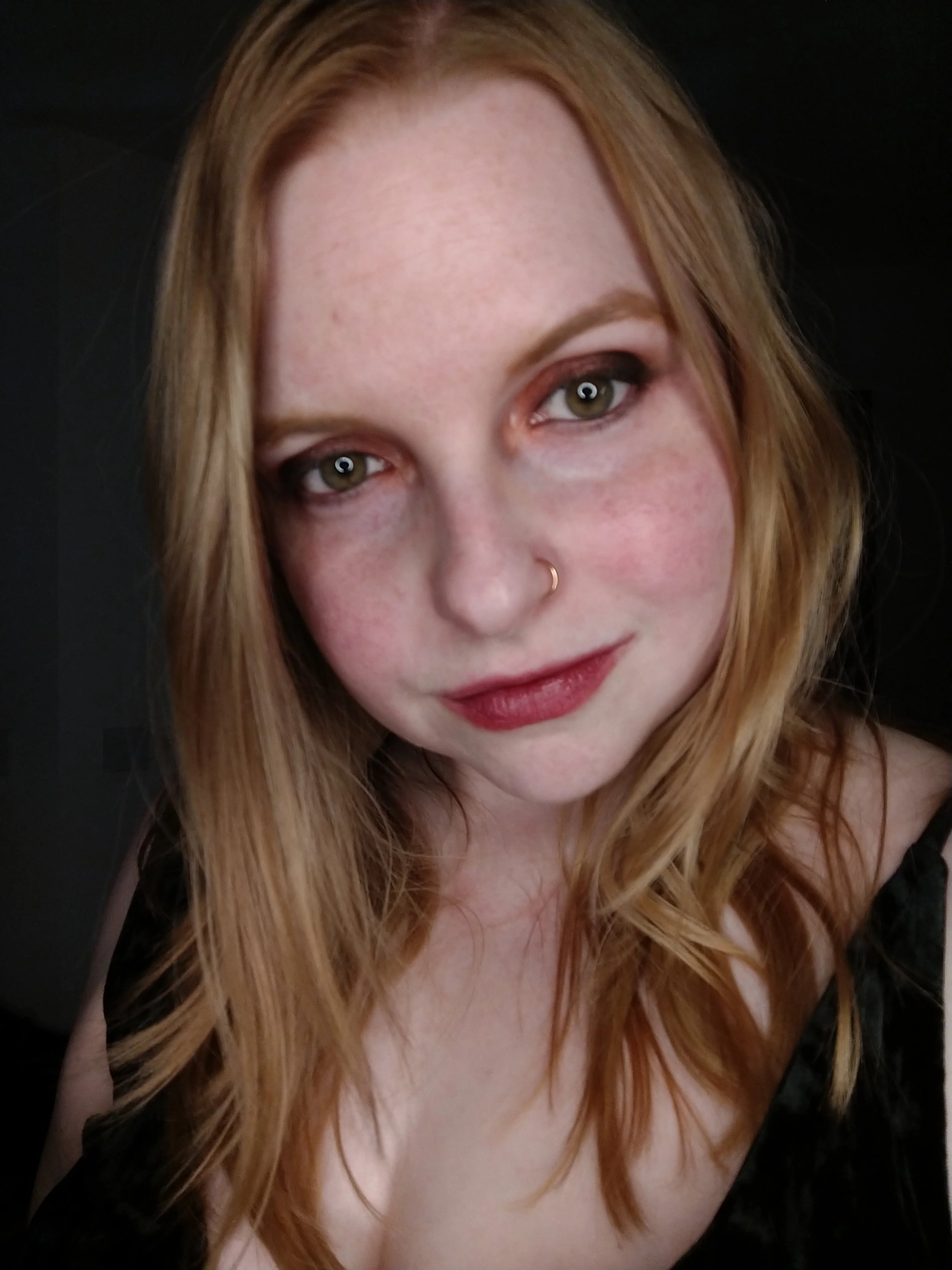 Close-up of a woman's face with long red hair, green eyes, a small nose ring, and wearing makeup, against a dark background.
