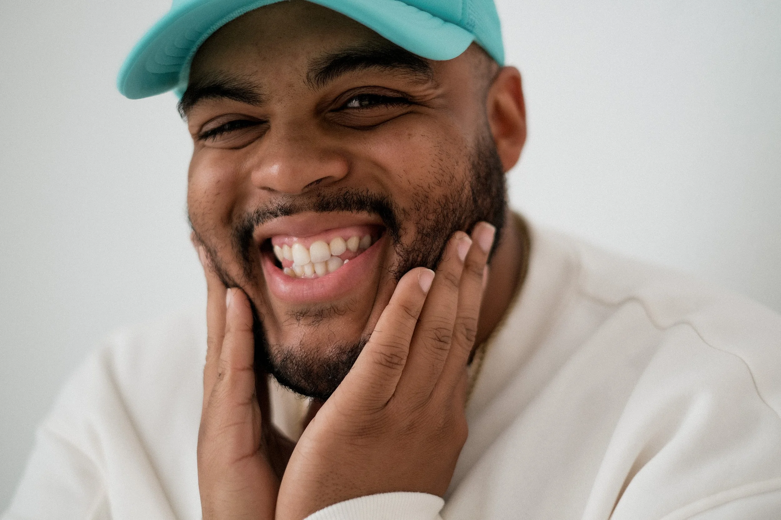 A man wearing a teal baseball cap smiling and touching his beard with his hand.