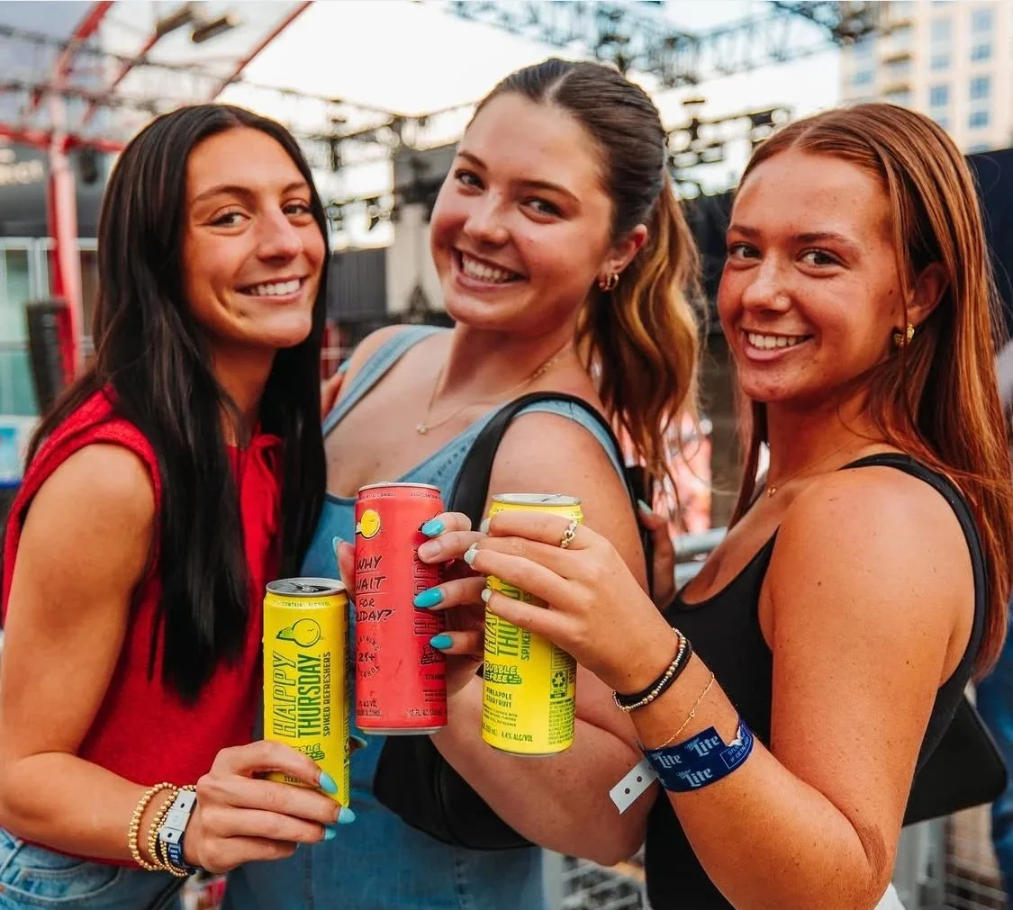 Three women at an outdoor event, smiling and holding cans of beverages, with a cityscape and stage in the background.