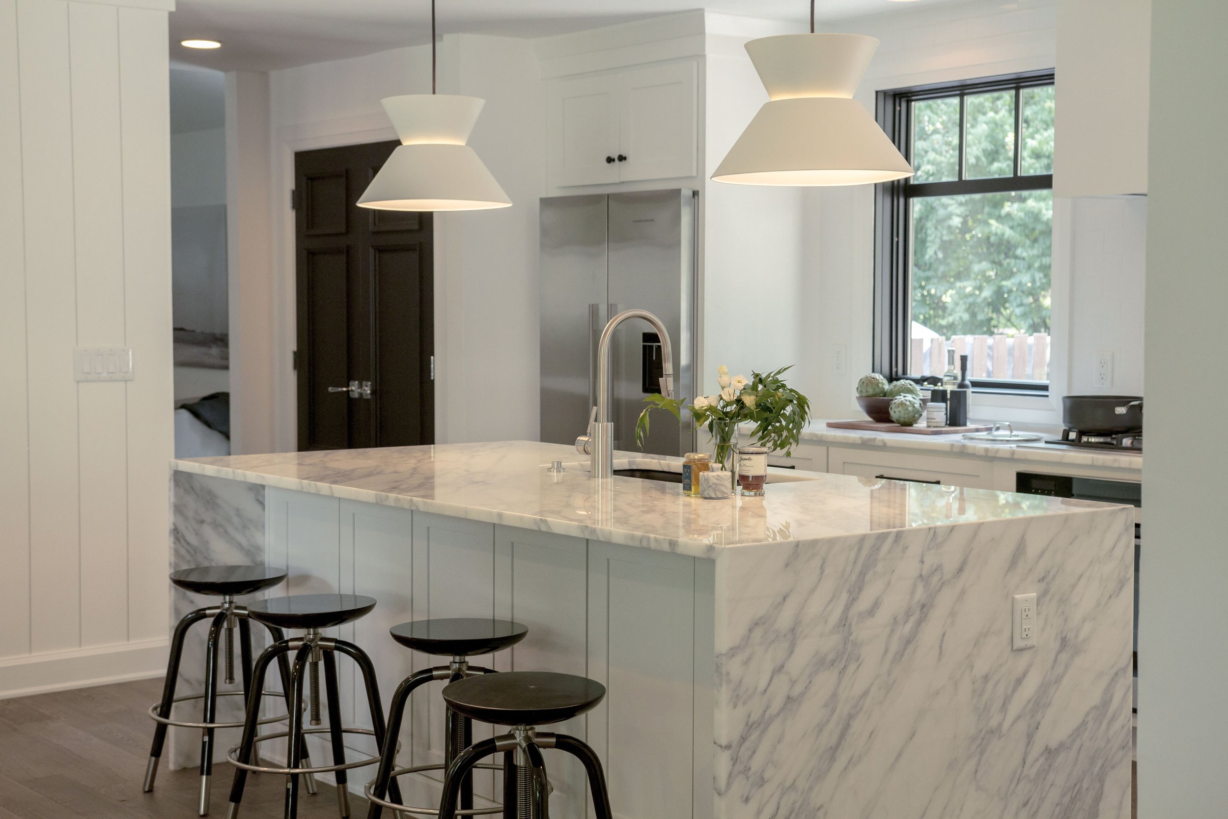 A modern kitchen with a marble island, three black bar stools, hanging pendant lights, a stainless steel refrigerator, and a window with a view of greenery outside.