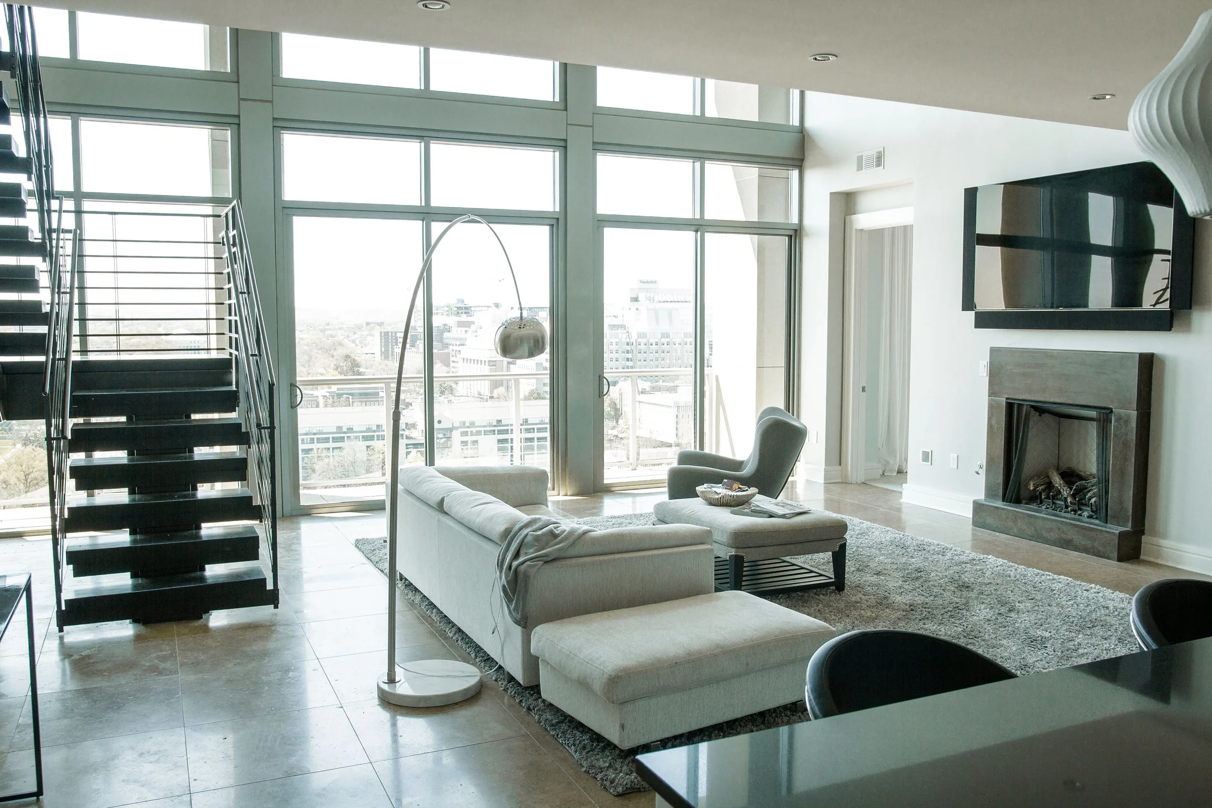 Modern living room with large windows, white couch, armchair, fireplace, mounted TV, and sliding glass doors leading to balcony.