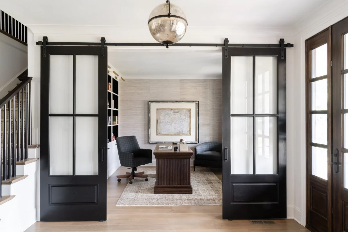 Home office with sliding barn doors, a wooden desk, black chair, black armchair, a framed map on the wall, bookshelf, and natural light from windowed door.