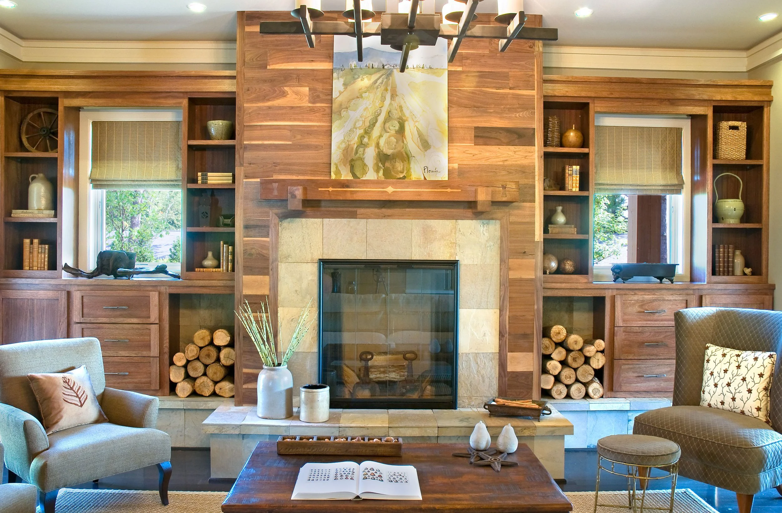 Living room with a central fireplace surrounded by wooden shelving, decorated with vases, books, and decorative objects. Two windows with blinds flanking the fireplace, and furniture including armchairs and a coffee table with decorative items.