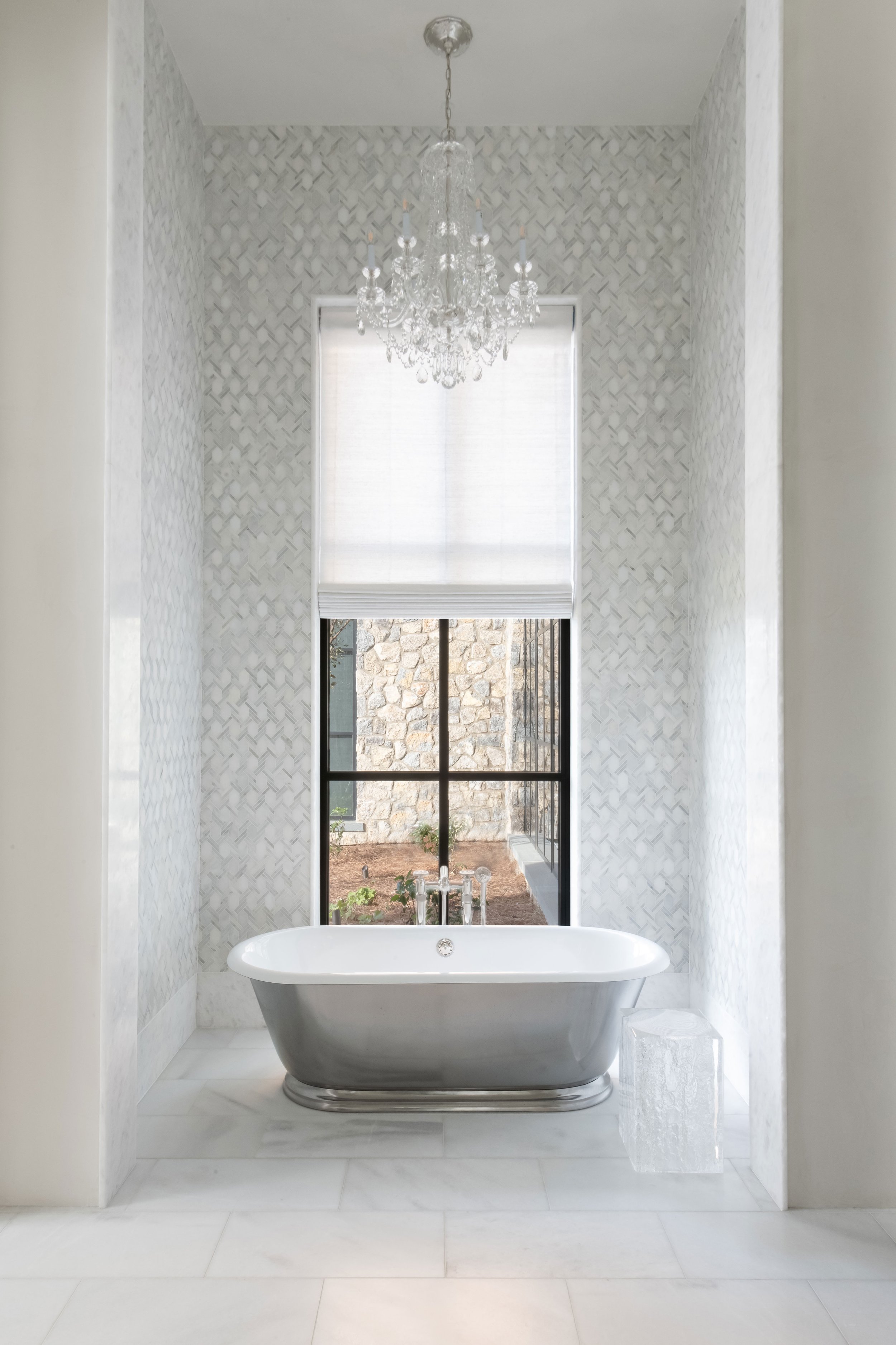 Elegant bathroom with freestanding bathtub in front of large window, chandelier hanging from ceiling, and textured walls.