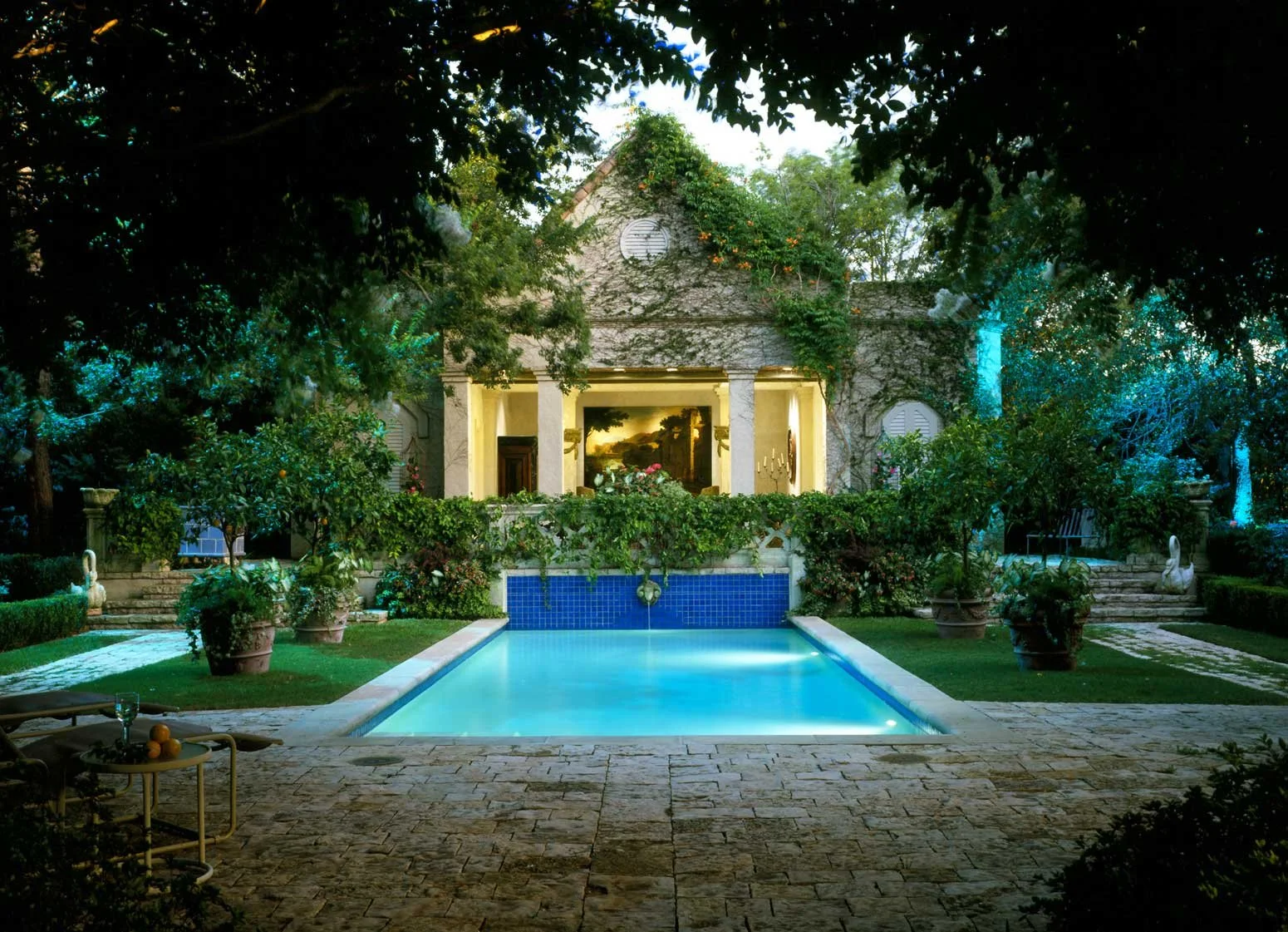A backyard with a swimming pool, surrounded by lush greenery and potted plants. A house with columns and a large window is in the background, illuminated at dusk.