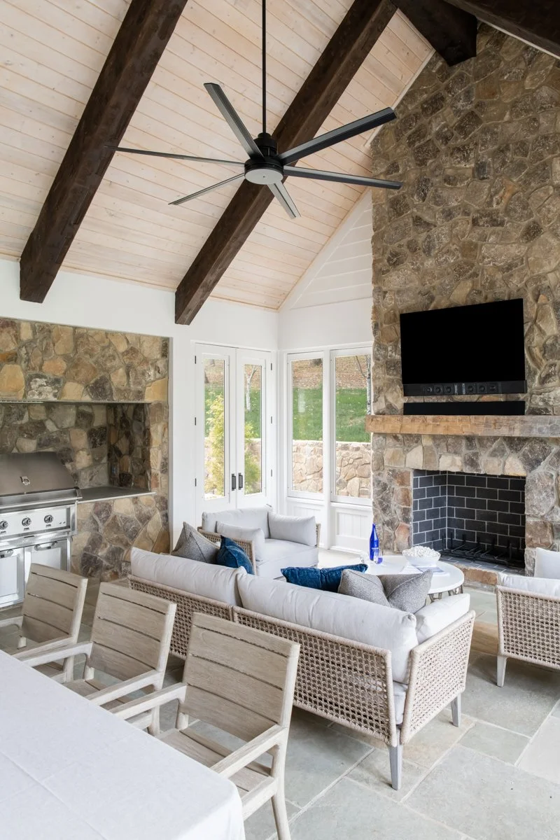 A living room with a vaulted ceiling, exposed wooden beams, and a stone fireplace with a mounted TV. There are large windows letting in natural light, and the room is furnished with a white sofa, a wicker chair, a coffee table, and a dining table wit