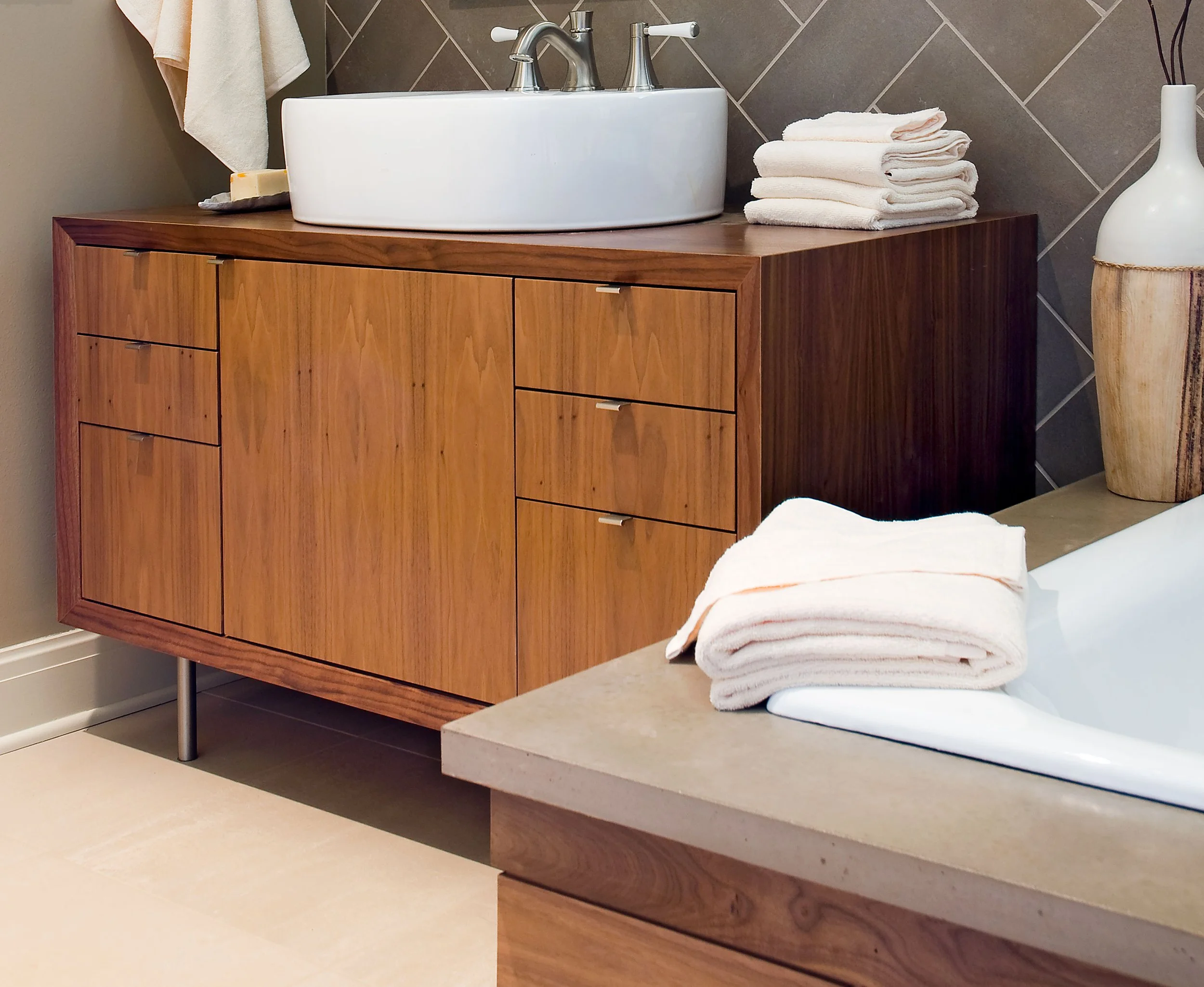 A bathroom vanity with a white vessel sink on top, folded white towels, and decorative items, with a bathtub nearby.