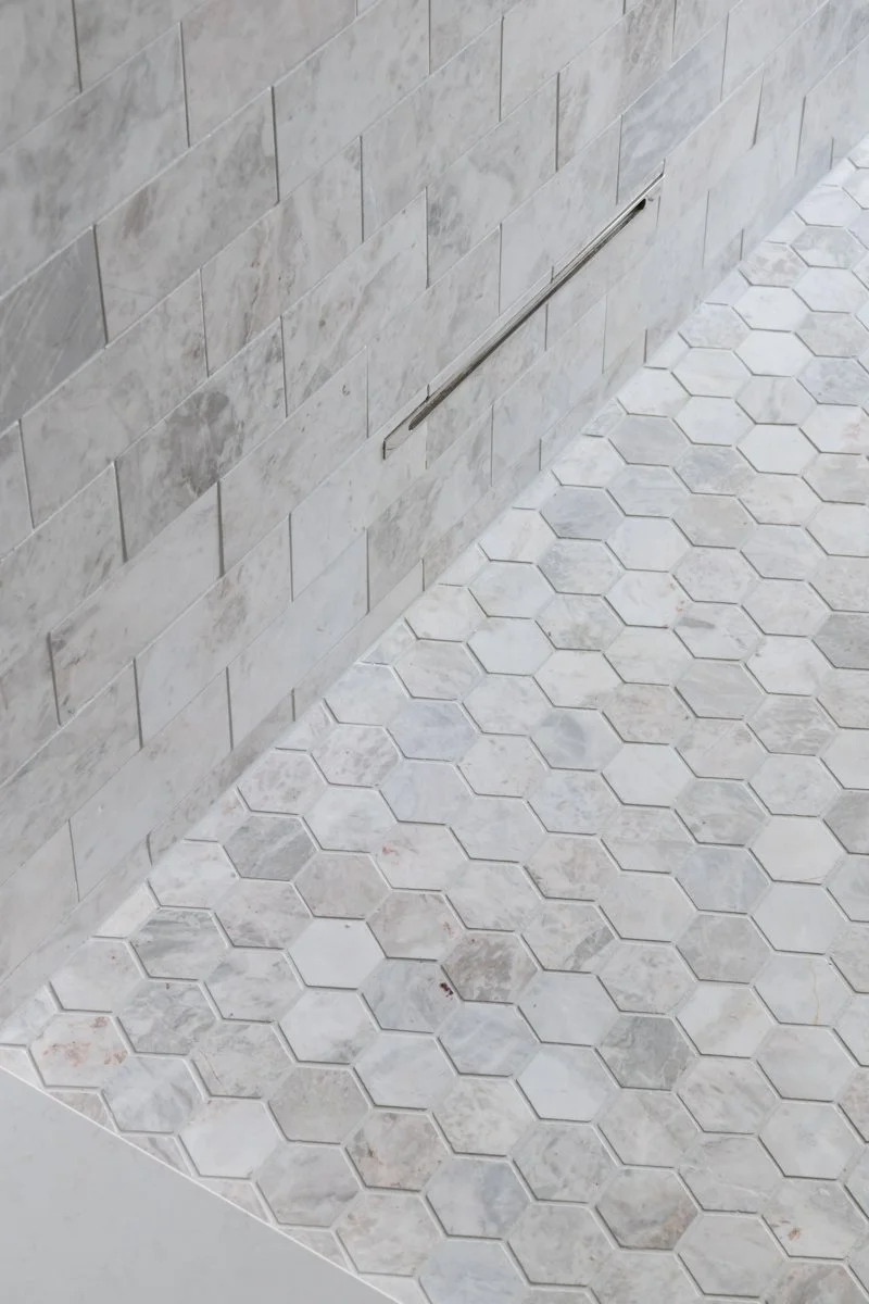 Close-up of a gray hexagonal tile floor with a partial inlaid tile border and a wall with light gray rectangular tiles, featuring a metal handrail.
