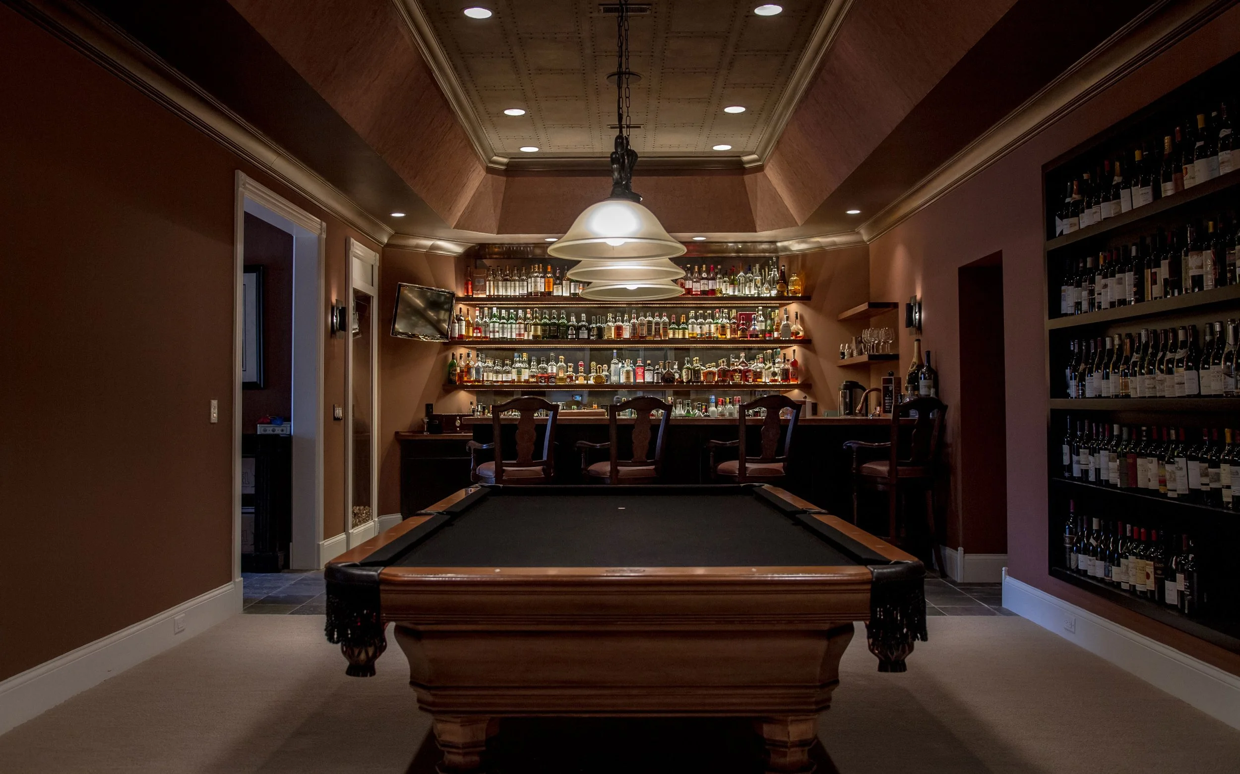 A luxurious basement game room with a pool table and a bar with shelves of liquor bottles in the background.