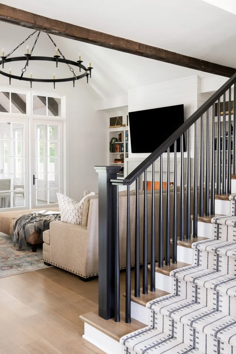 Living room with a staircase, TV, bookshelf, beige armchair, sofa, chandelier, and large windows.
