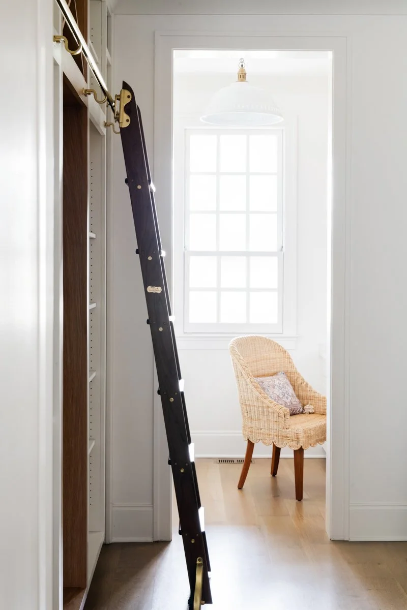 Interior of a bright room with a large window, a wicker chair with a cushion, and a ladder leaning against a built-in bookshelf or closet.