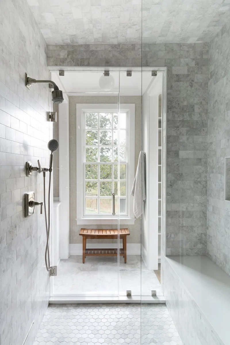 Bright bathroom with a walk-in shower, large window, wooden bench, white tiled walls, and gray tiled walls.