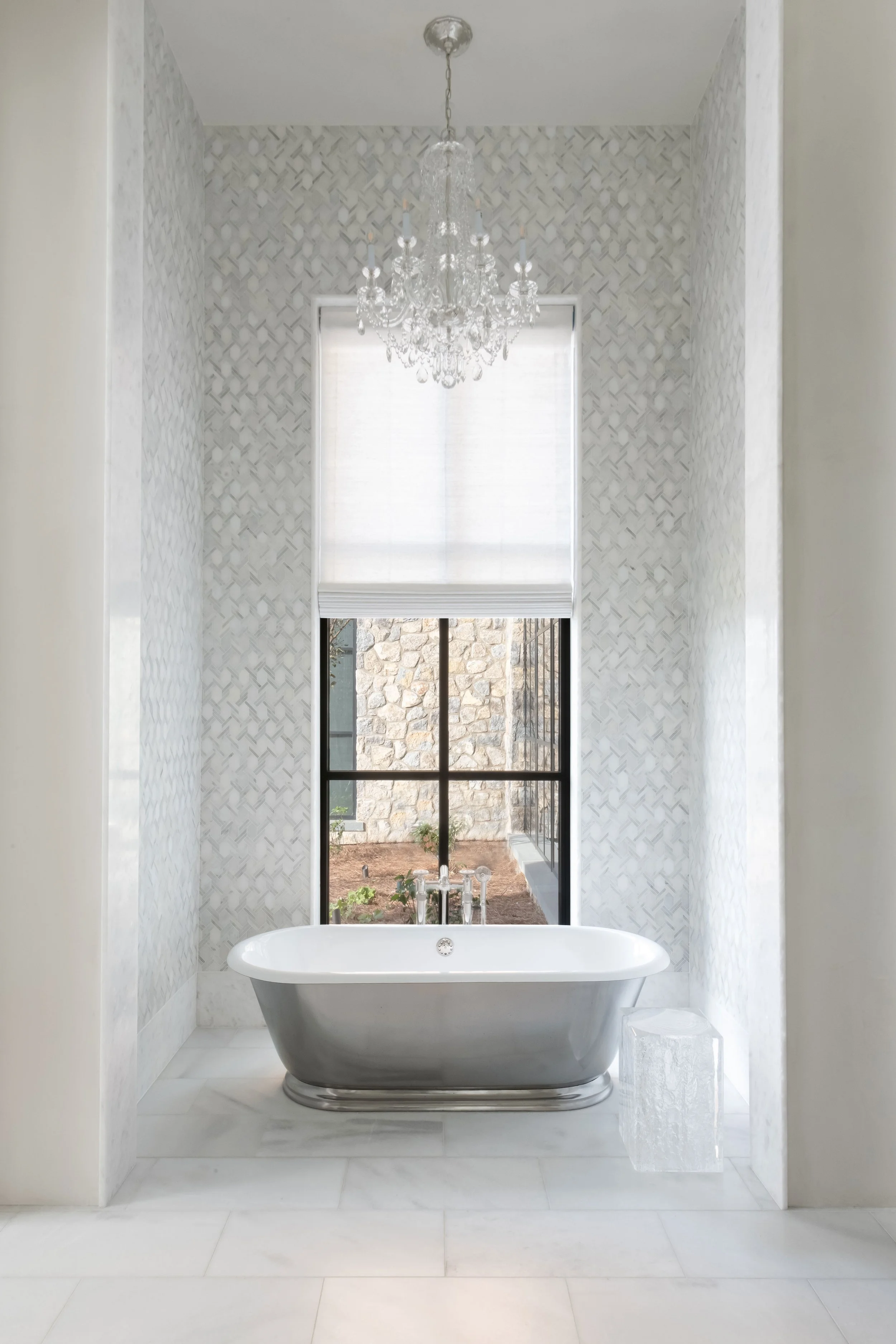 A luxurious bathroom features a freestanding bathtub beneath a large window, with a chandelier hanging overhead and a small side table beside the tub.