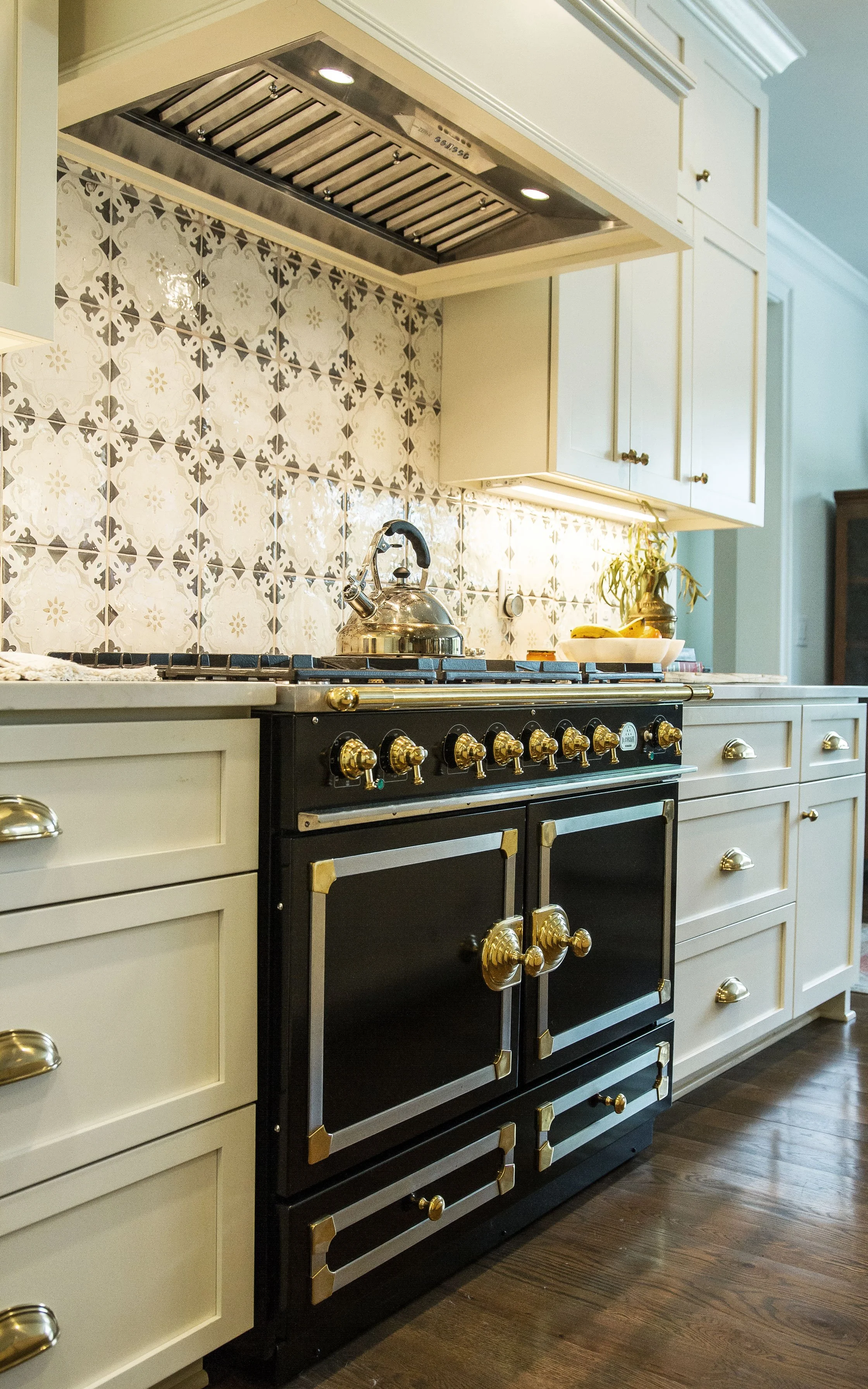 A kitchen with a black vintage-style stove with gold accents, white cabinets, a patterned tile backsplash, and a kettle on the stove.