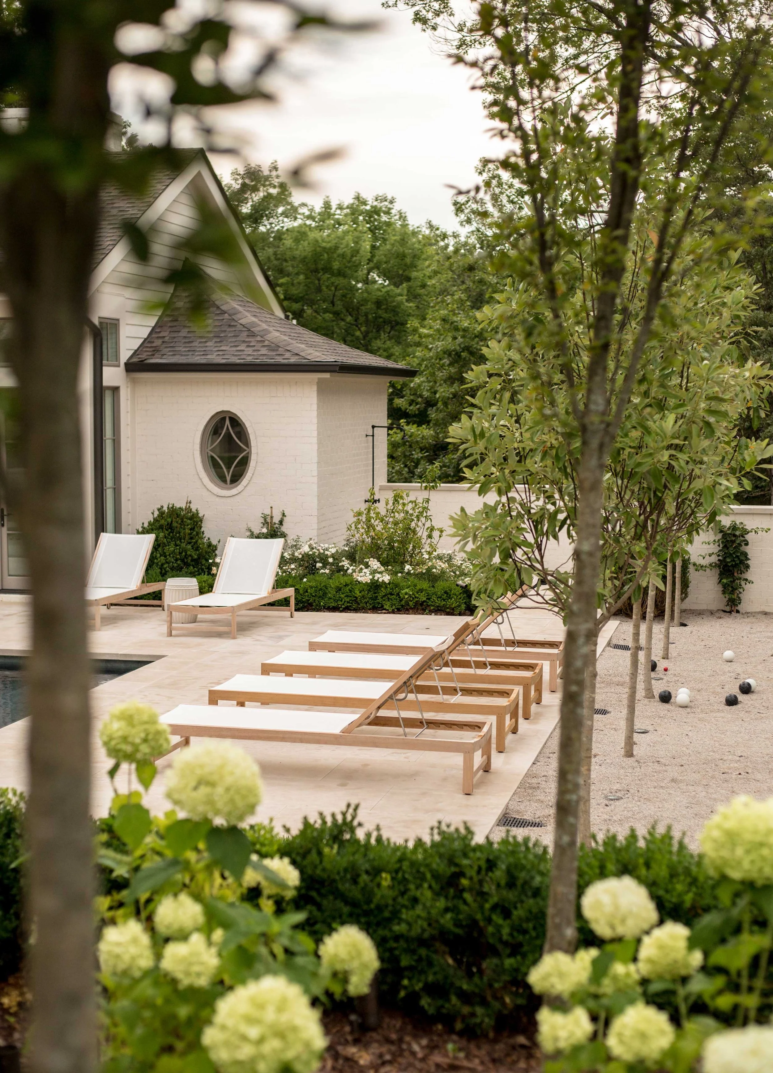 A backyard with a swimming pool, white lounge chairs, young trees, flowering bushes, and a light-colored house with a round window.