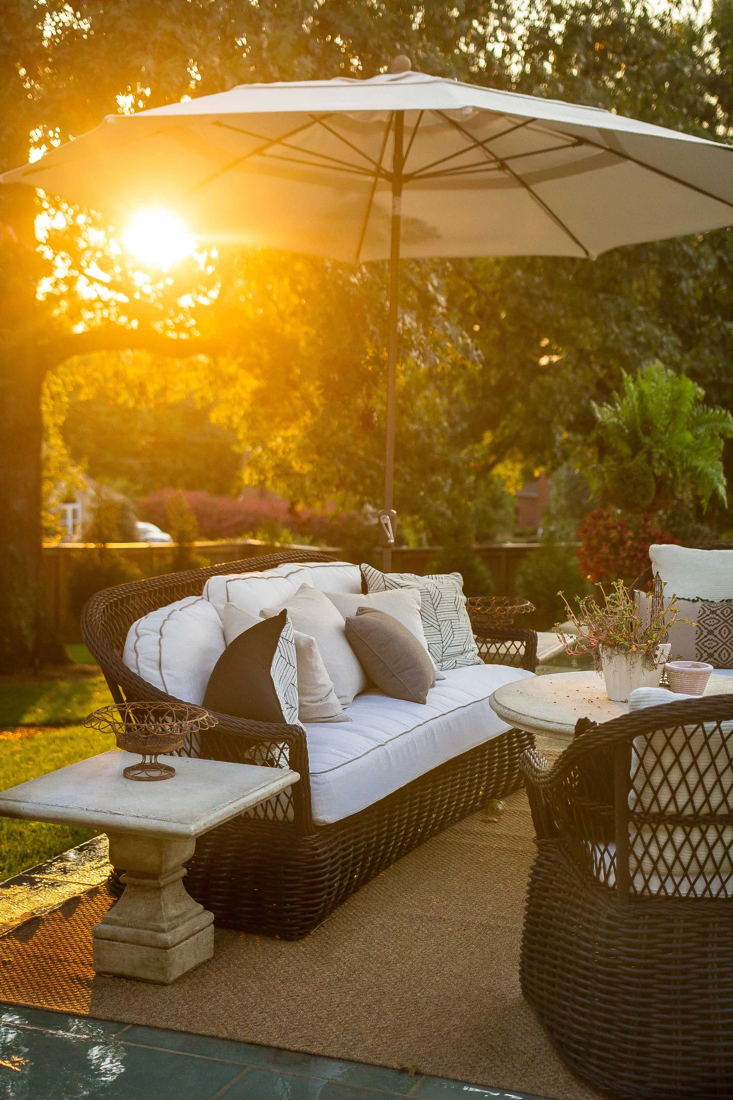 An outdoor patio scene during sunset, featuring a wicker sofa with white cushions and multiple throw pillows, a round stone table with a potted plant, a matching wicker chair, and a large white umbrella providing shade.