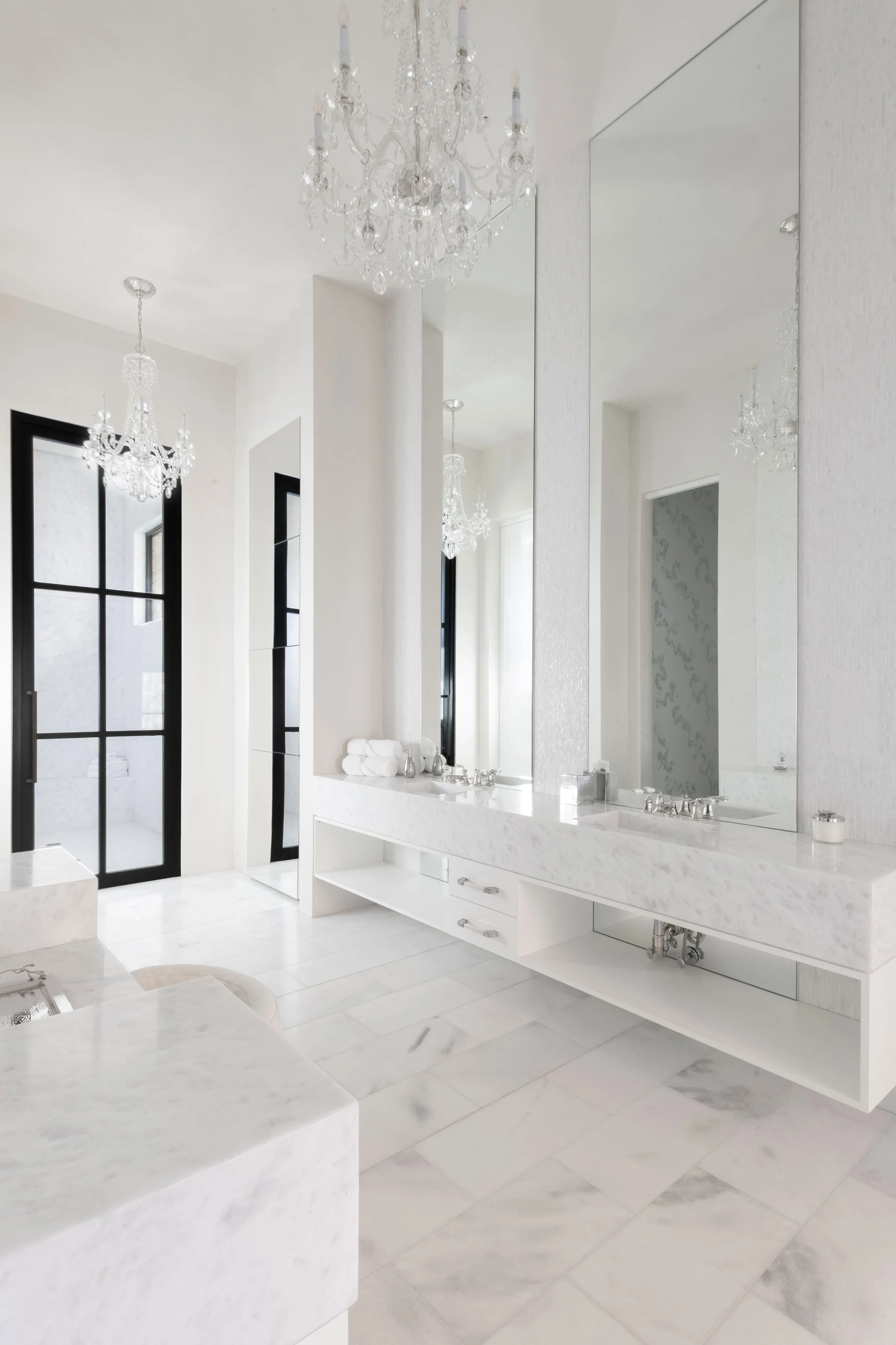 A luxurious modern bathroom with white marble countertops, a large mirror, black-framed windows, and elegant chandeliers.