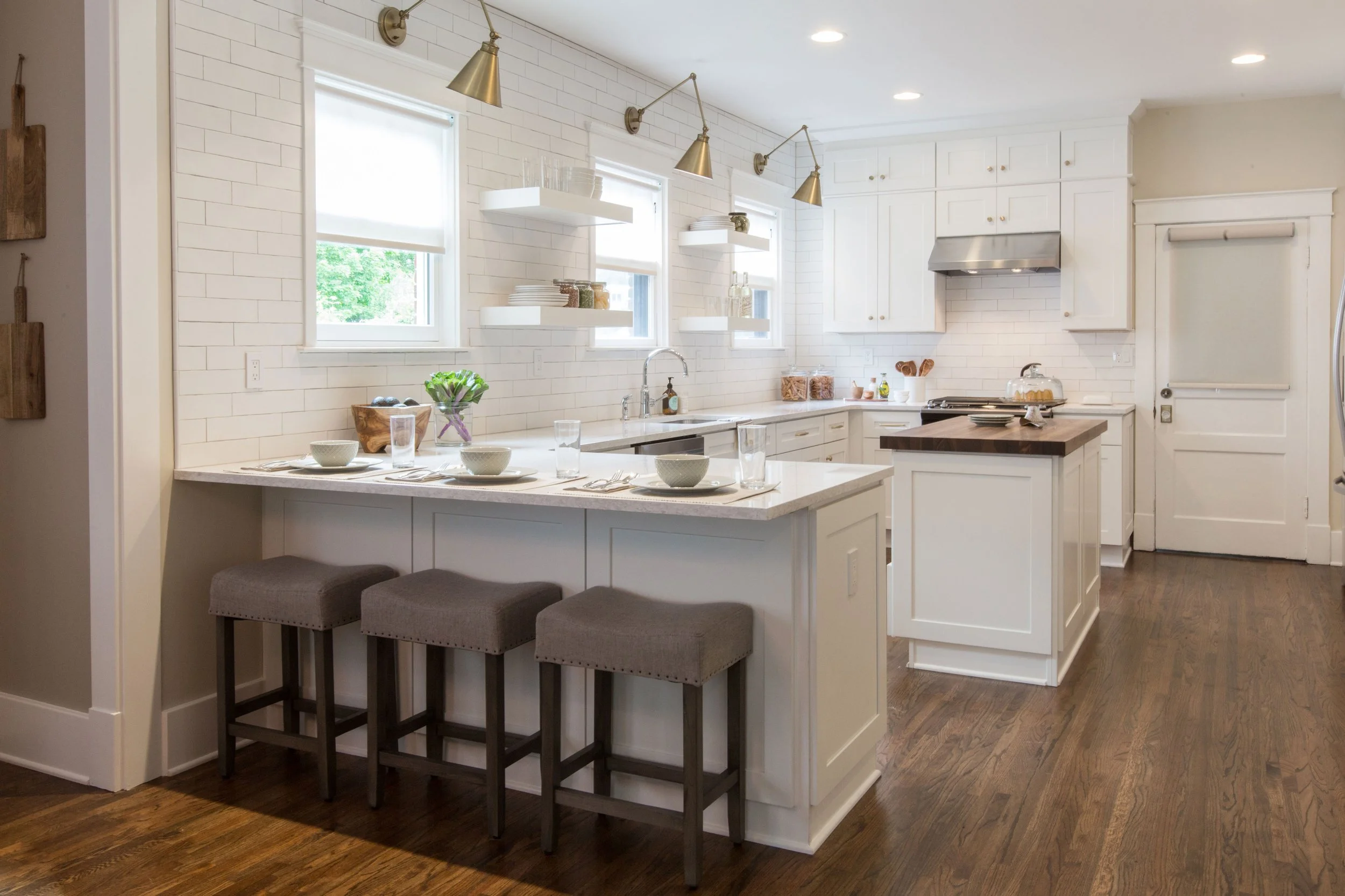 Modern white kitchen with island, three brown stools, three windows, open shelves, and wood floors.