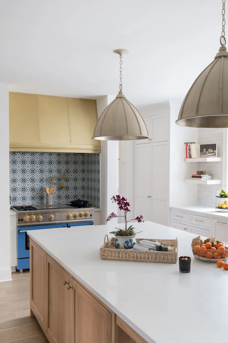 A modern kitchen with white cabinetry, a large white island, and two large pendant lights hanging above the island. The kitchen has a yellow range hood, patterned blue backsplash, and open shelves with books and decorative items. On the island, there