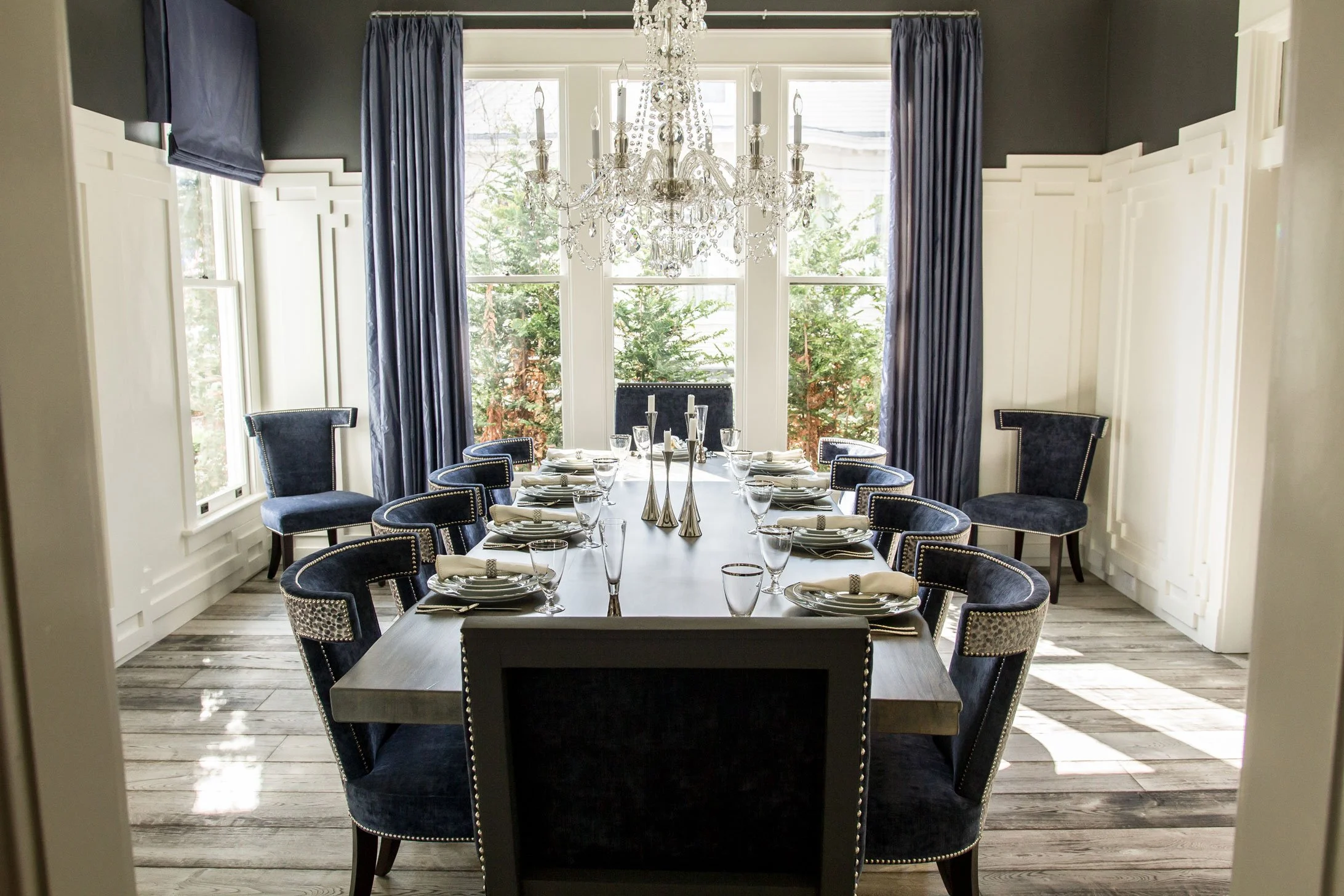 Elegant dining room with a long table set for a meal, surrounded by navy velvet chairs with metallic nailhead trim, large windows with dark curtains, and a crystal chandelier hanging above.