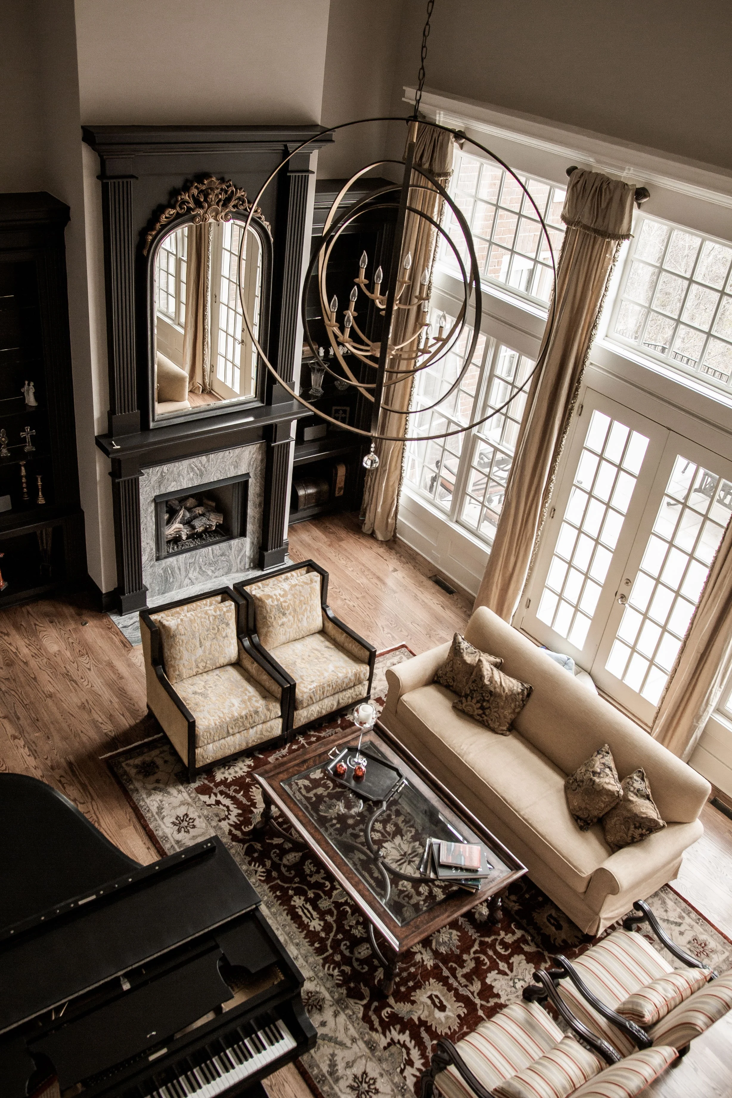 Overhead view of a living room with a fireplace, a chandelier, a large window, a sofa, two armchairs, a coffee table, and a piano.