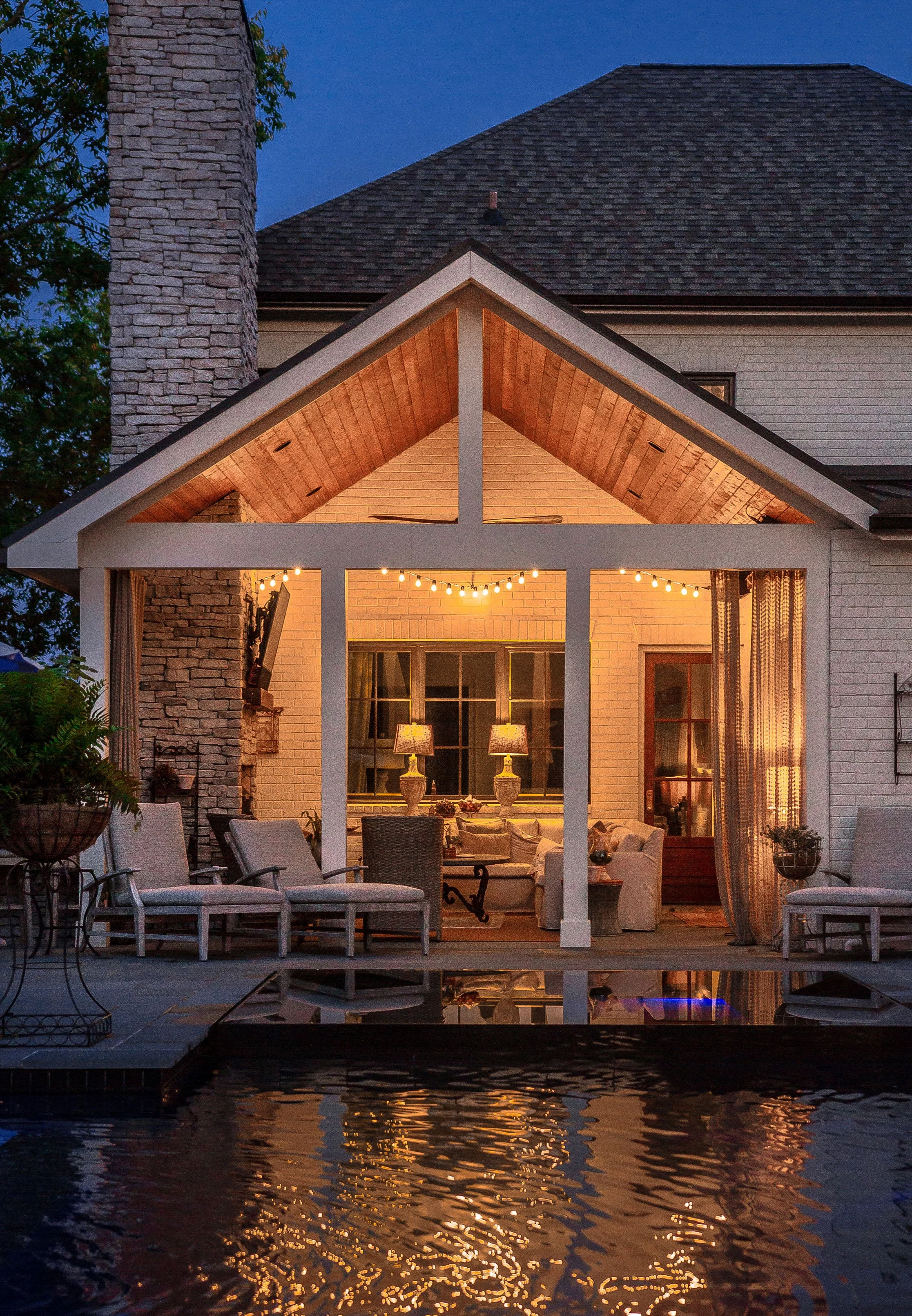 Back porch with lounge chairs, tables with lamps, string lights, and a view of a swimming pool reflecting the lit porch area