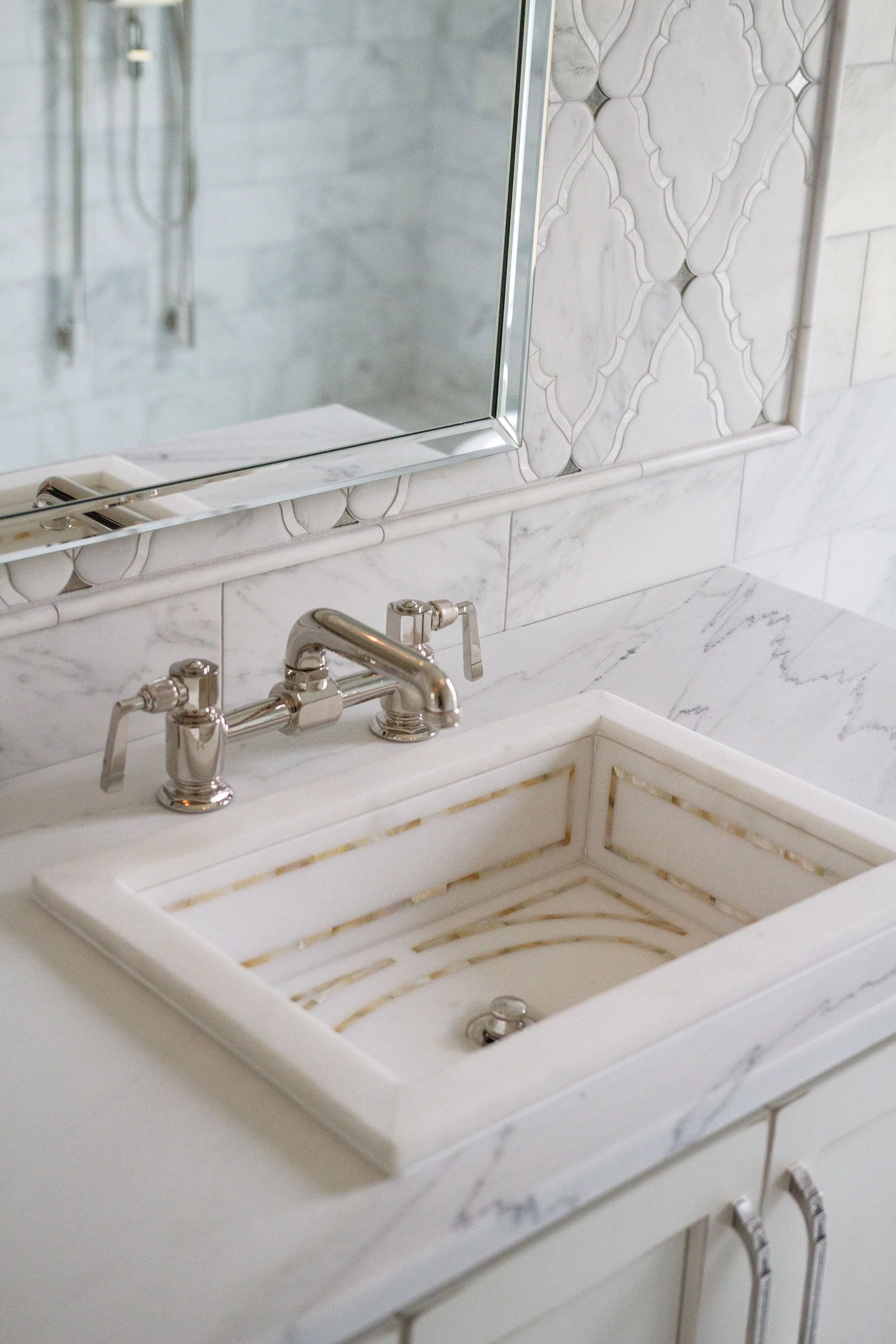 Close-up of a white marble bathroom sink with a reflective silver faucet and a large mirror above. The surrounding wall features intricate marble tile patterns.