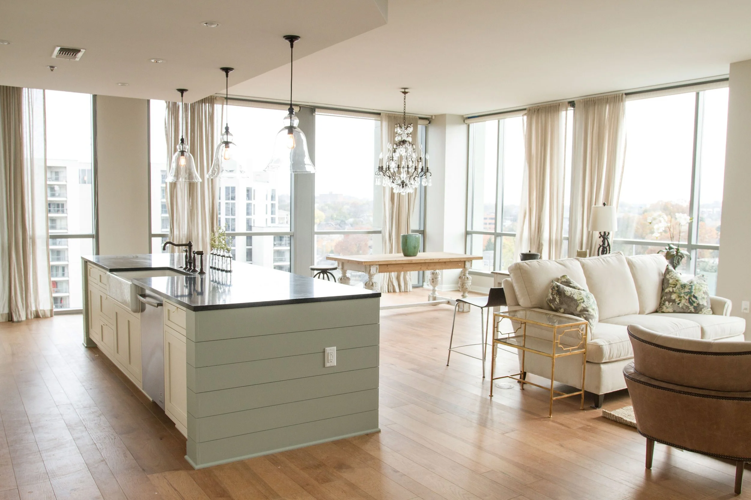 Bright modern living room with large windows, white walls, hardwood floors, a beige sofa with floral pillows, a gold side table, a black armchair, a white kitchen island with a black countertop, pendant lights, and a chandelier.