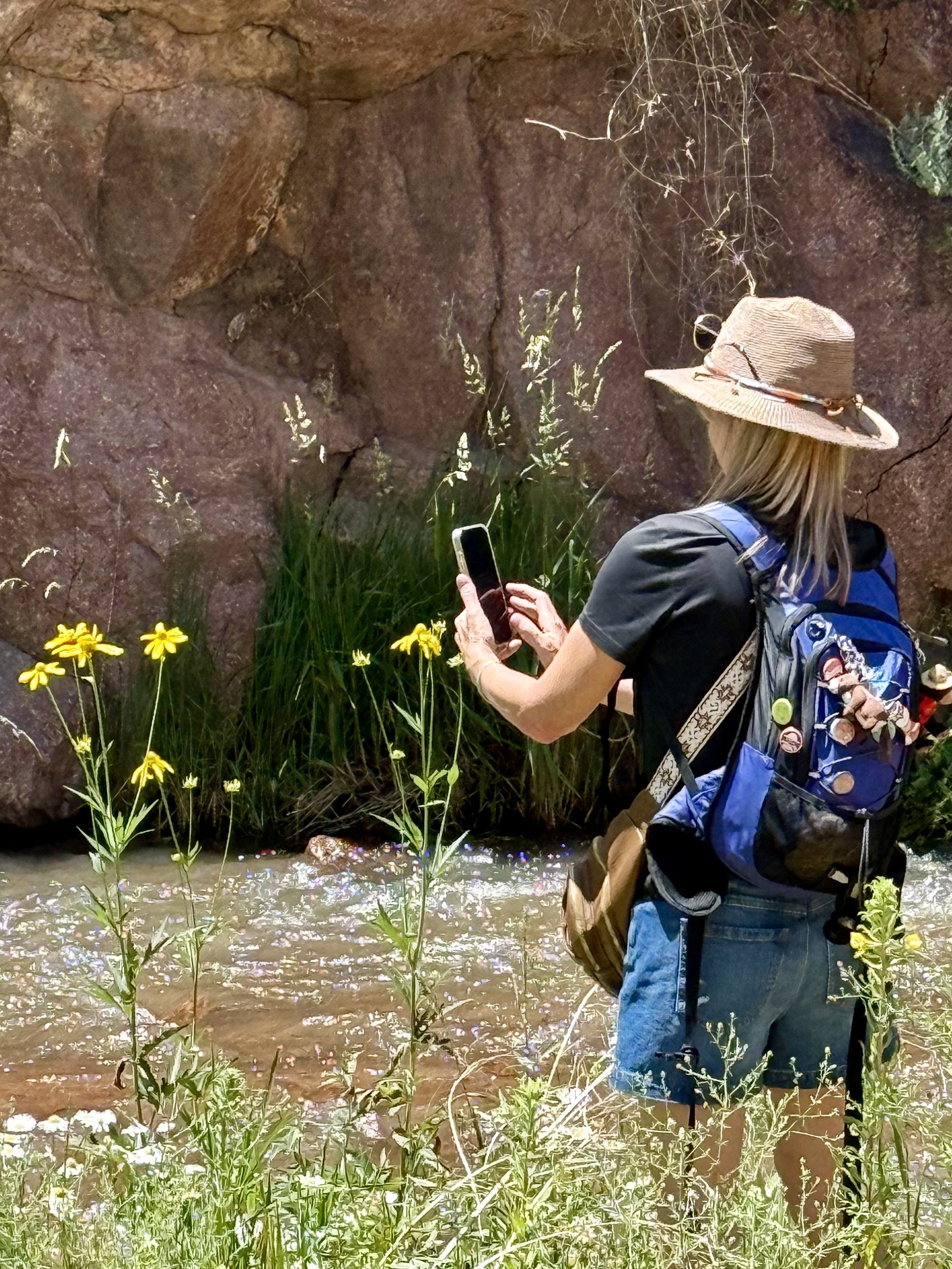 A woman wearing a wide-brimmed hat and a backpack standing by a rocky creek, taking a photo with her smartphone.