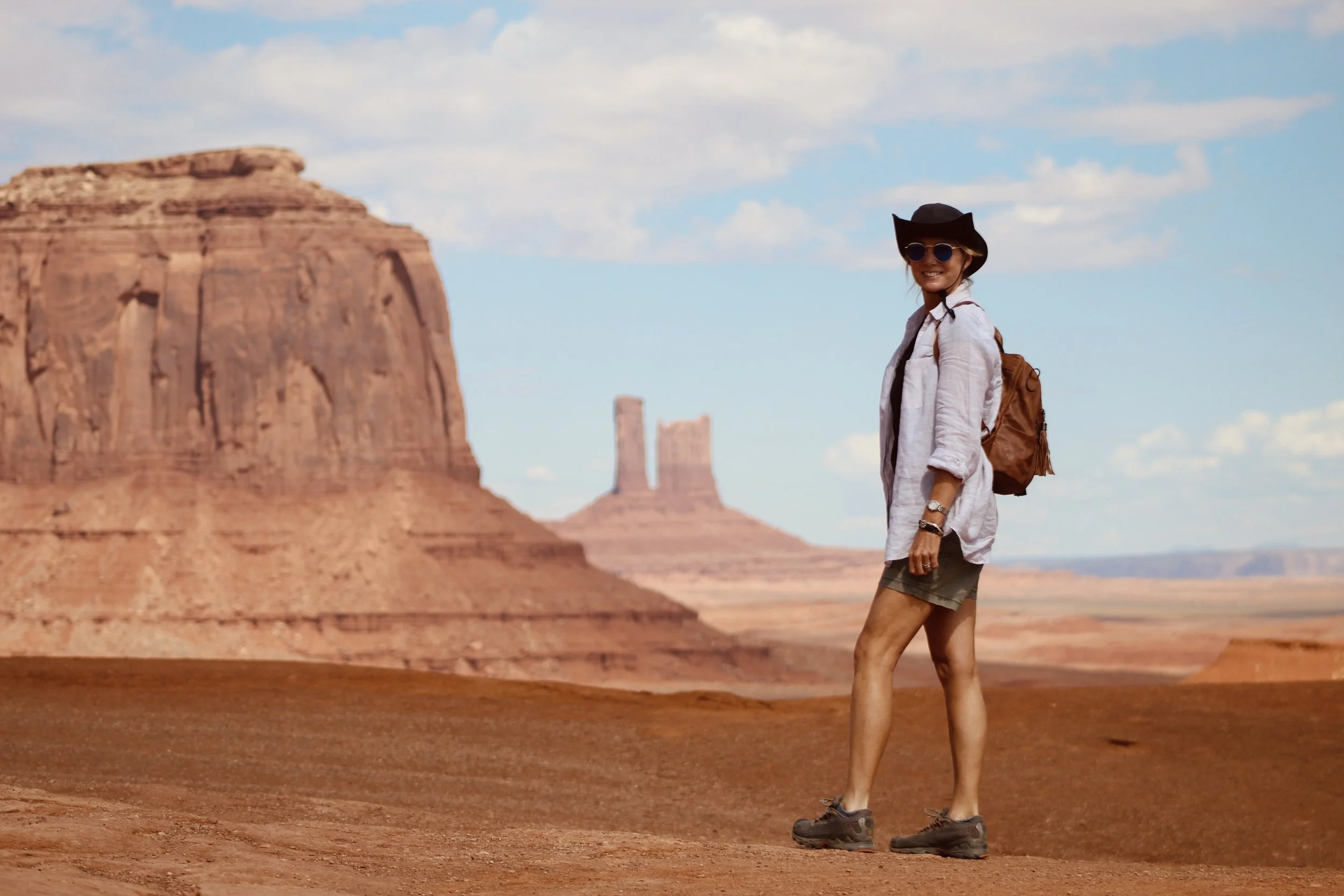 A woman wearing a black hat, sunglasses, a white shirt, and hiking gear standing in a desert landscape with large rock formations and blue sky.