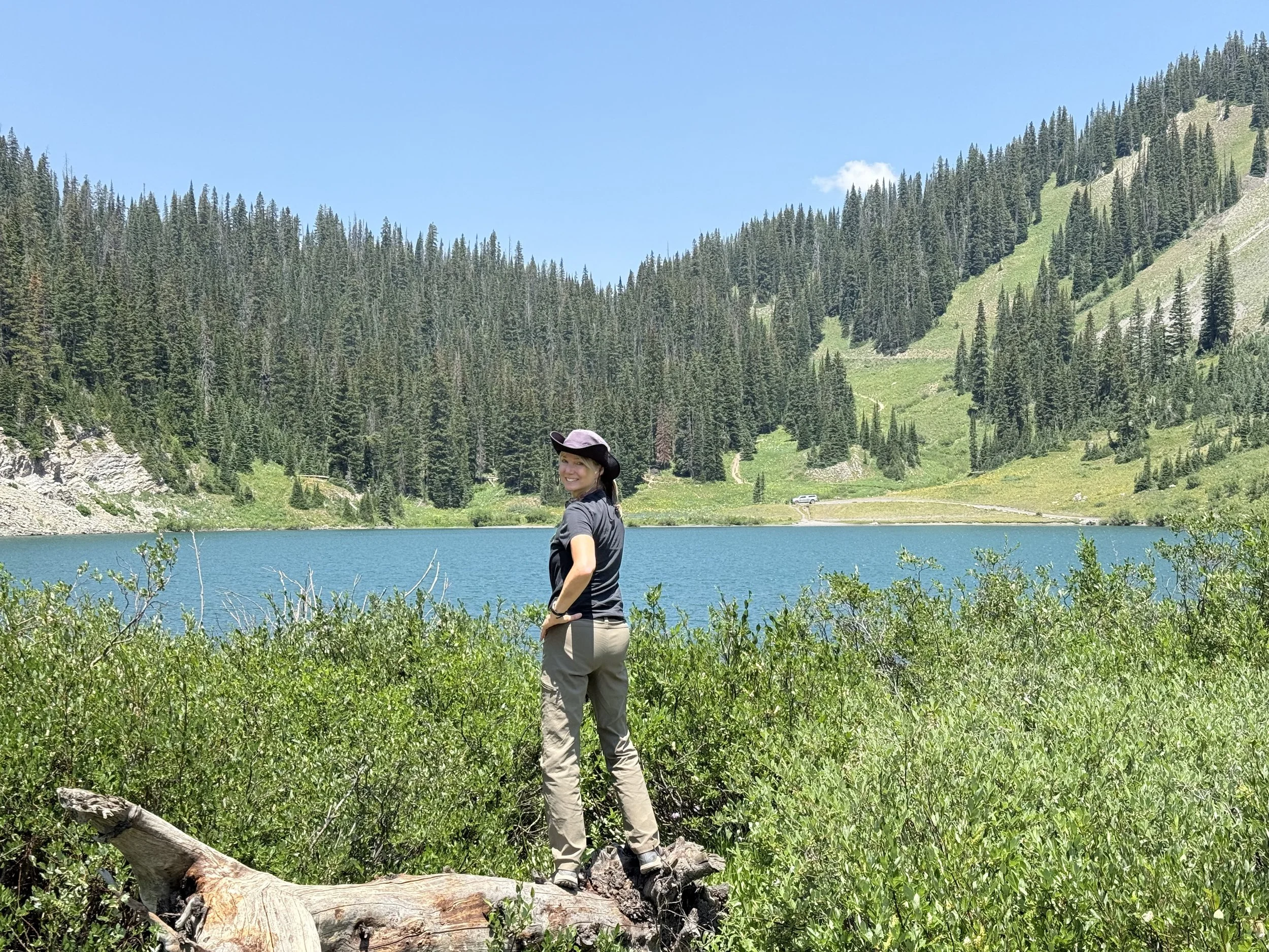 A woman standing on a fallen tree near a lake with a forested mountain in the background. She is wearing a hat and outdoor clothing, smiling, facing sideways.