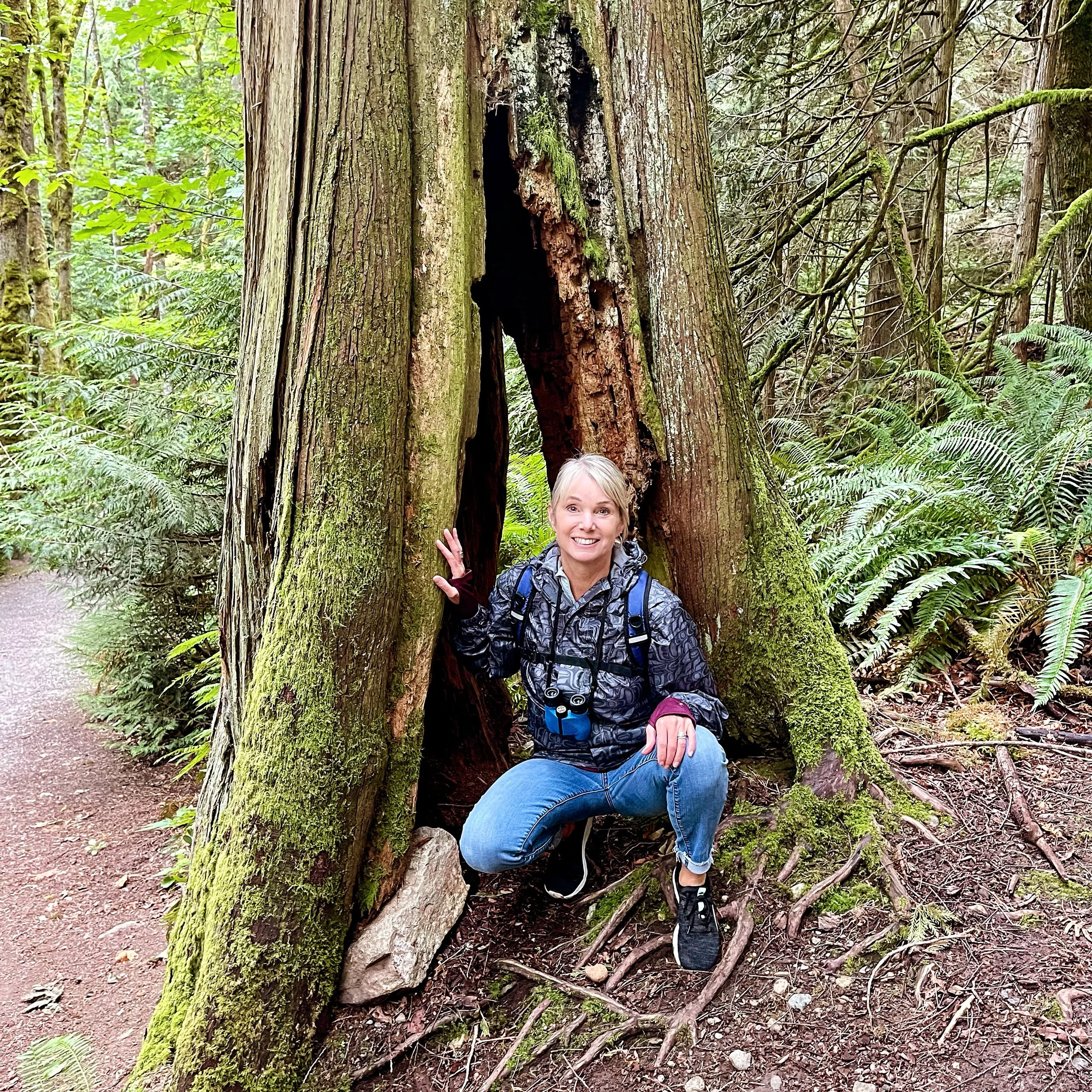 A woman smiling and making a peace sign pose next to a large felled tree trunk covered in moss in a lush, green forest.