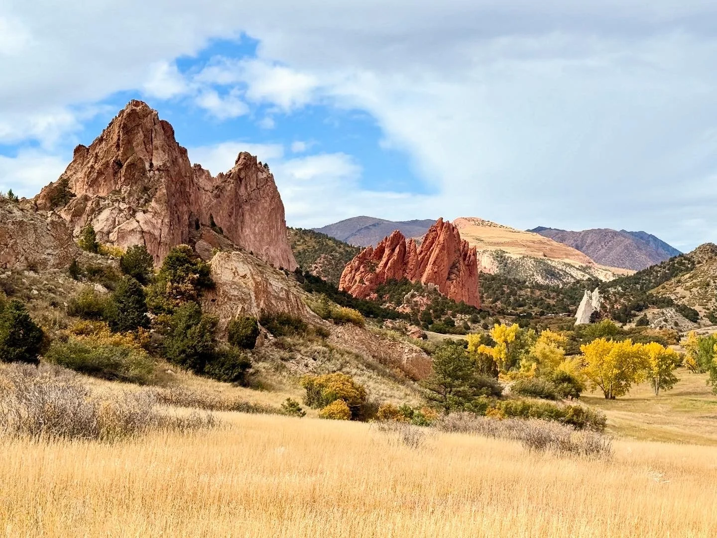 A scenic landscape featuring tall, reddish and pinkish rocky mountains against a partly cloudy sky, with a grassy field and trees in fall colors in the foreground.