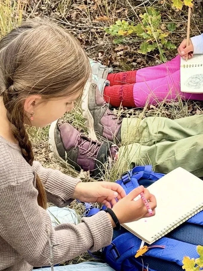 A young girl with braided hair is sitting outdoors on the grass, drawing or writing in a notebook with a pink pen. She is wearing a brown sweater, and her backpack is nearby. Another child, partially visible, is sitting nearby, holding a drawing pad.