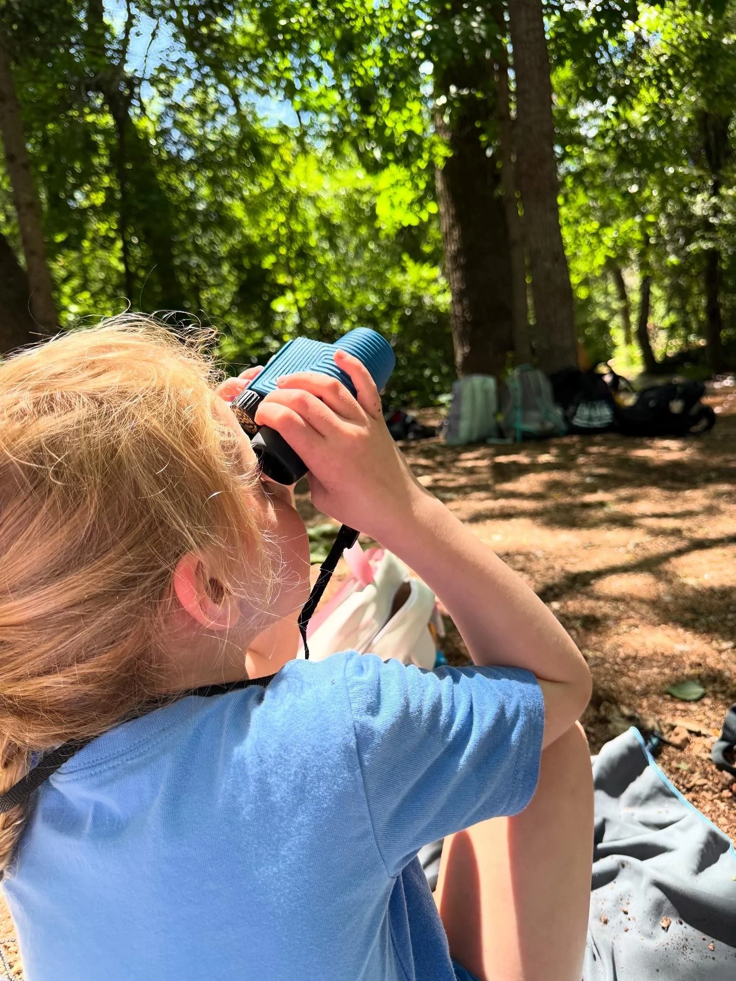 Girl with red hair looking through binoculars in a wooded area with backpacks nearby