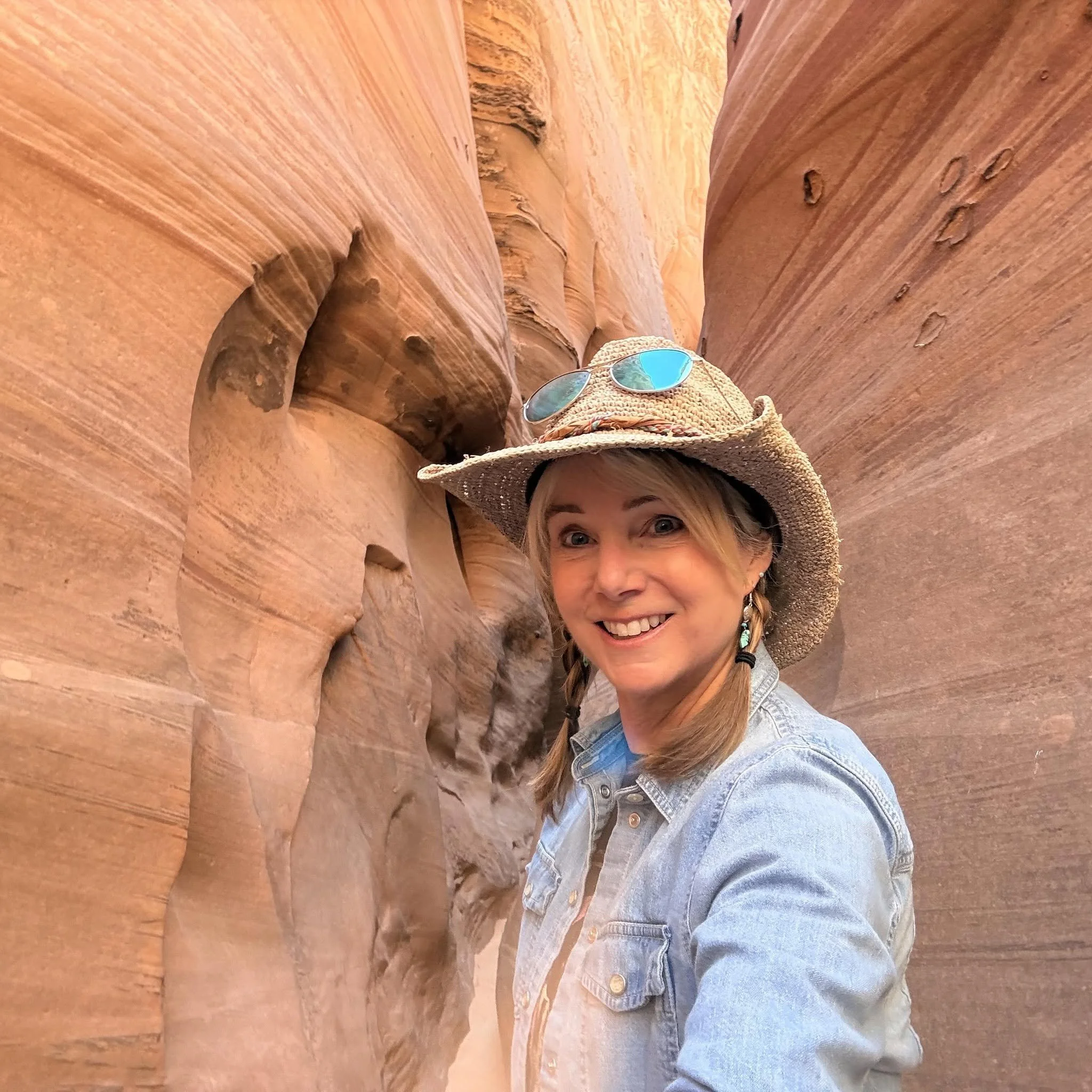 A woman with blonde hair and wearing a straw hat with sunglasses and a denim jacket, leaning against a rock formation in a canyon.