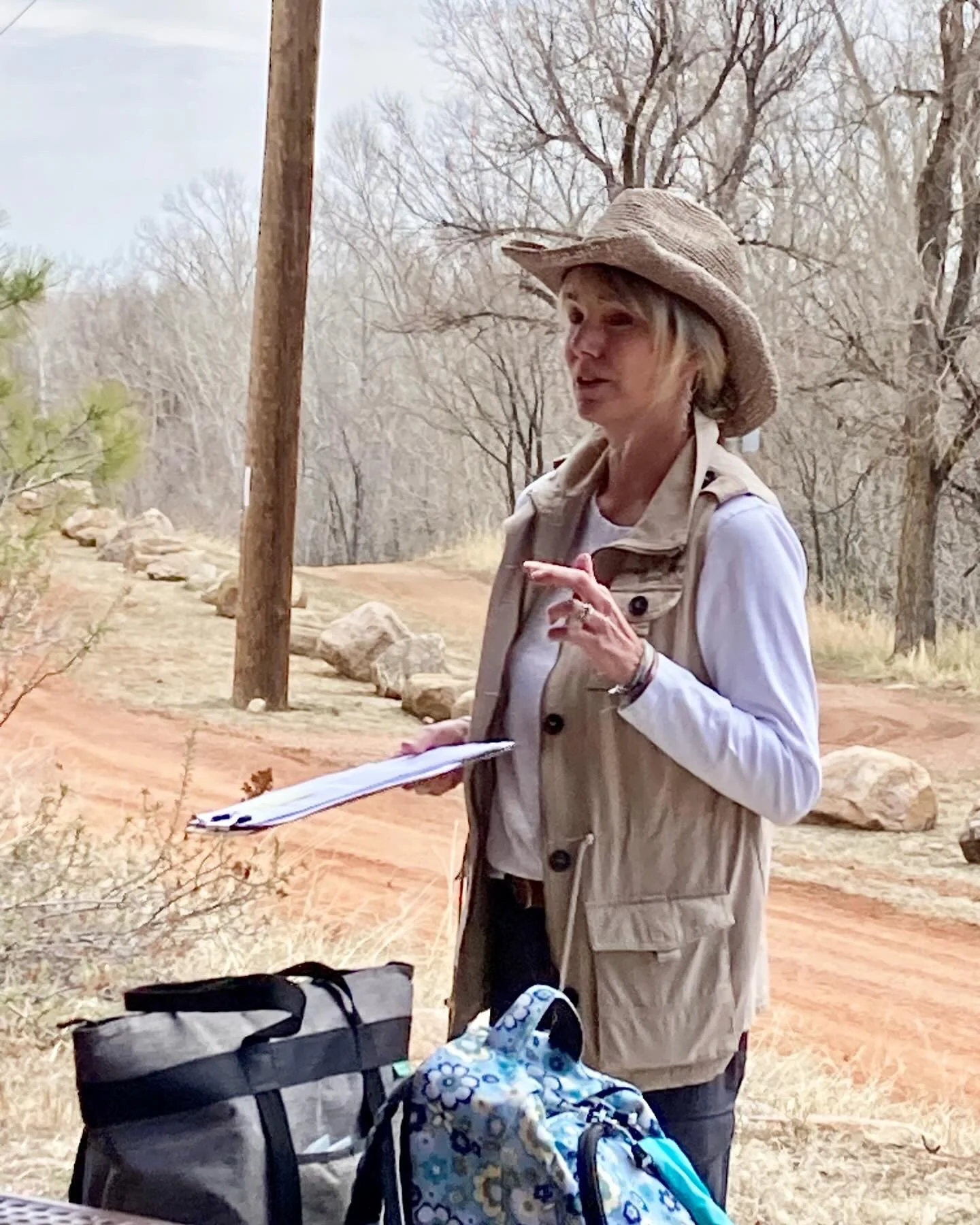 A woman wearing a large straw hat and tan vest is outdoors on a dirt path, holding a clipboard and speaking. She has a backpack and a smaller blue floral-patterned bag beside her, with trees and rocks in the background.
