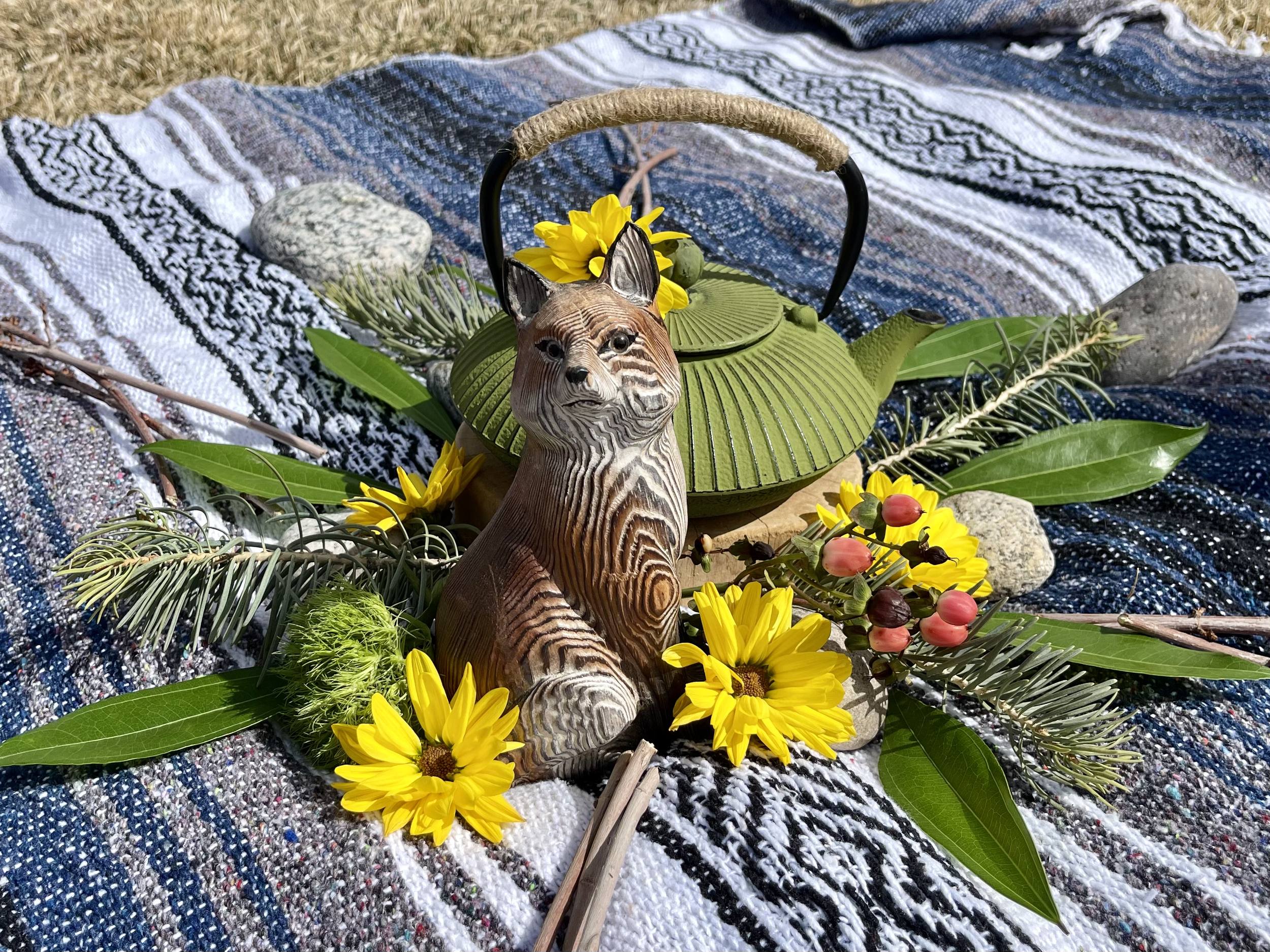 Decorative arrangement with a green teapot, a ceramic fox figure, yellow flowers, various green leaves and branches, and rocks on a striped fabric outdoors.
