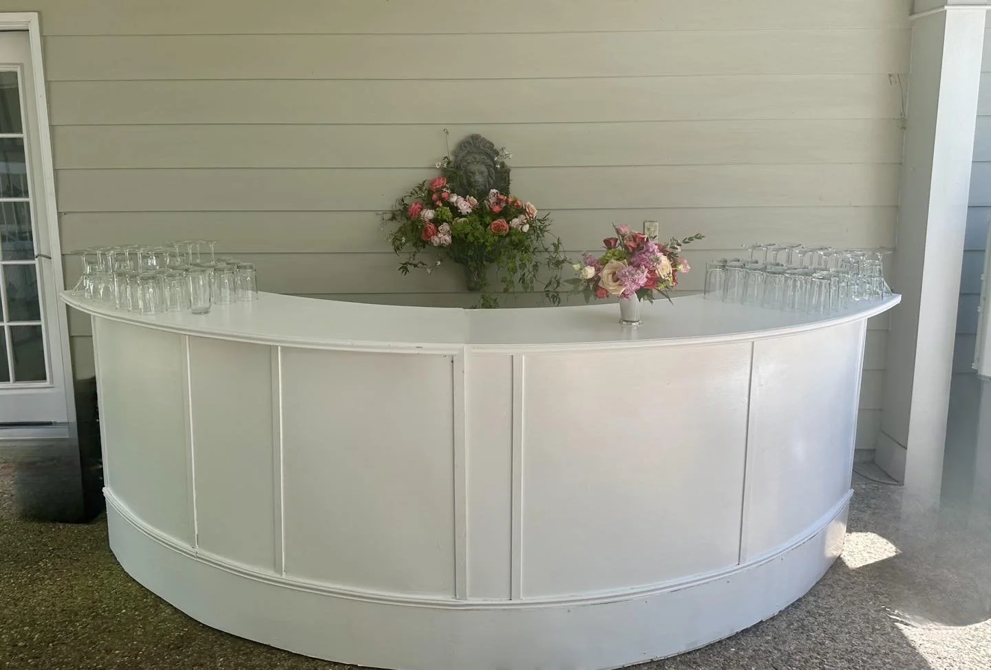 White patio bar with flower arrangements and glasses on top, set against a light-colored wooden wall.