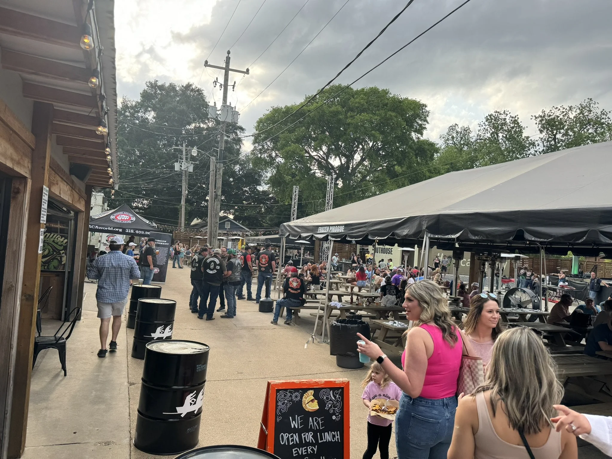 People gathered at an outdoor event with tents and picnic tables, some standing and others sitting, under a cloudy sky, with trees in the background.