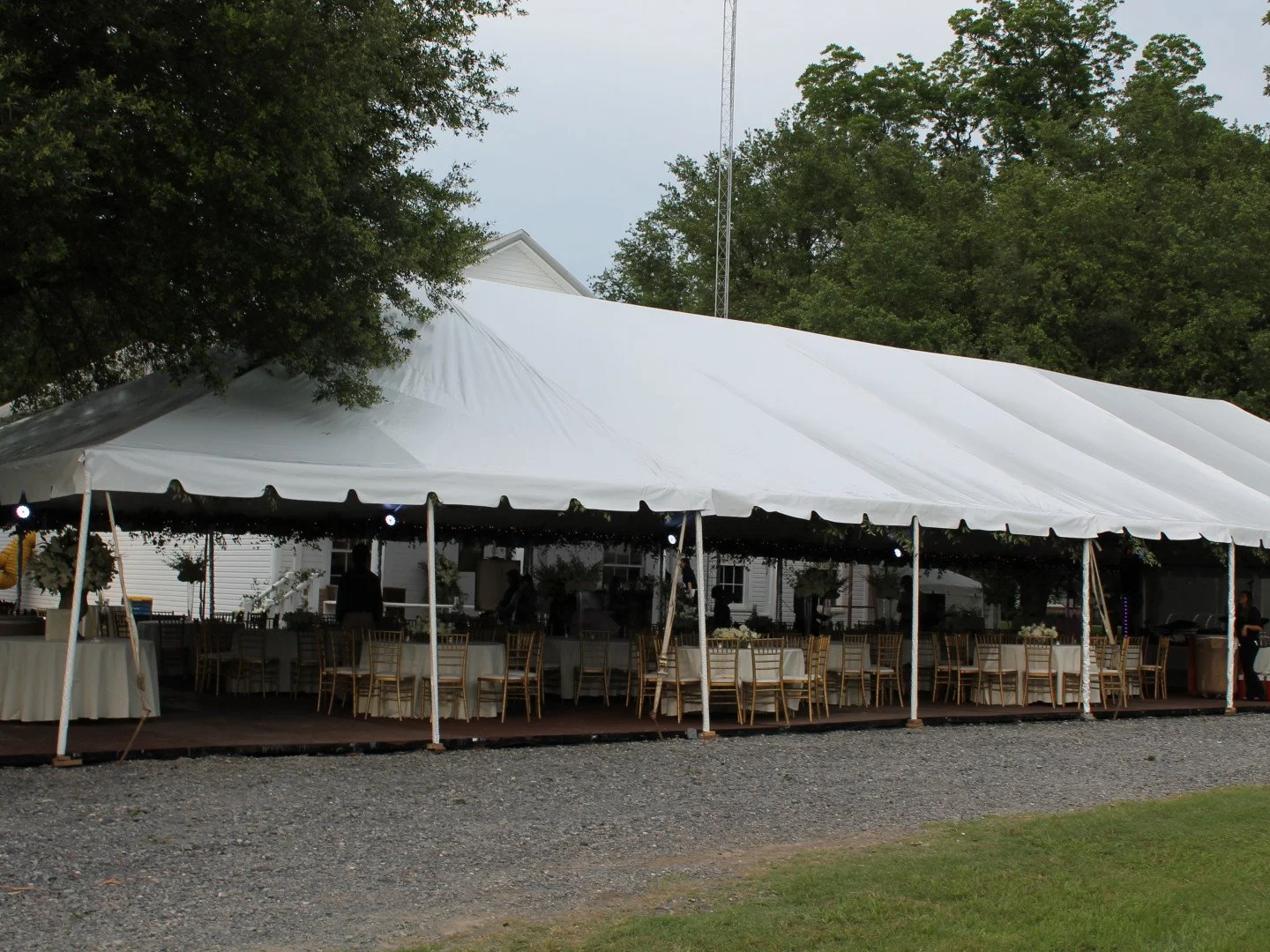 Large white event tent with gold chairs and tables underneath, set up outdoors on a gravel surface, surrounded by trees and a grassy area.