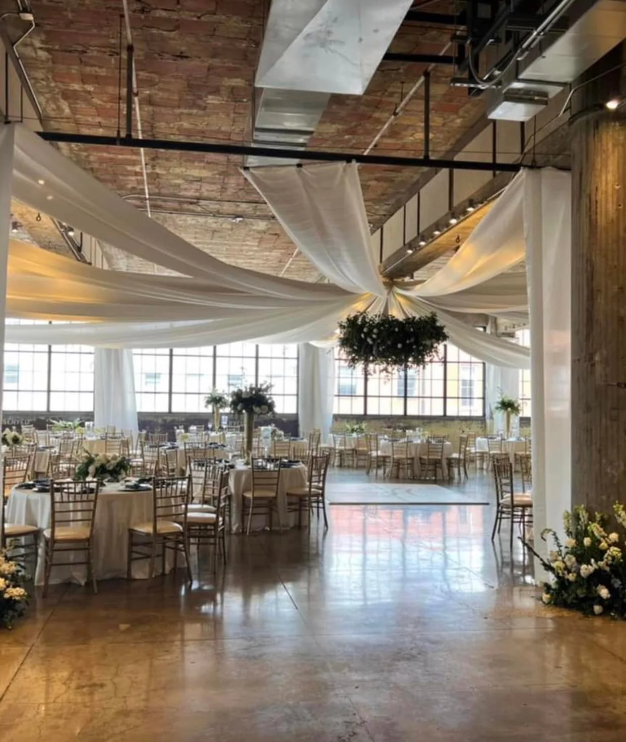 Elegant event space with round tables covered in white tablecloths, decorated with floral centerpieces, surrounded by wooden chairs, with white drapery hanging from the ceiling and a large floral arrangement hanging in the center, bright natural ligh