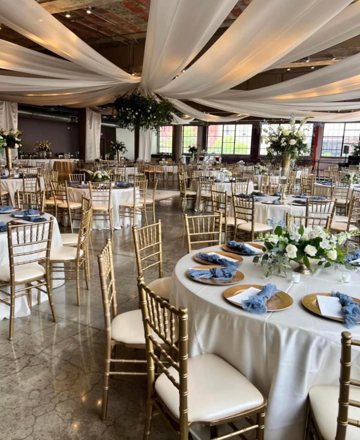Elegant event space decorated with draped white fabric on the ceiling, large floral centerpieces on round tables, and gold chairs with white cushions, set up for a celebration or wedding.
