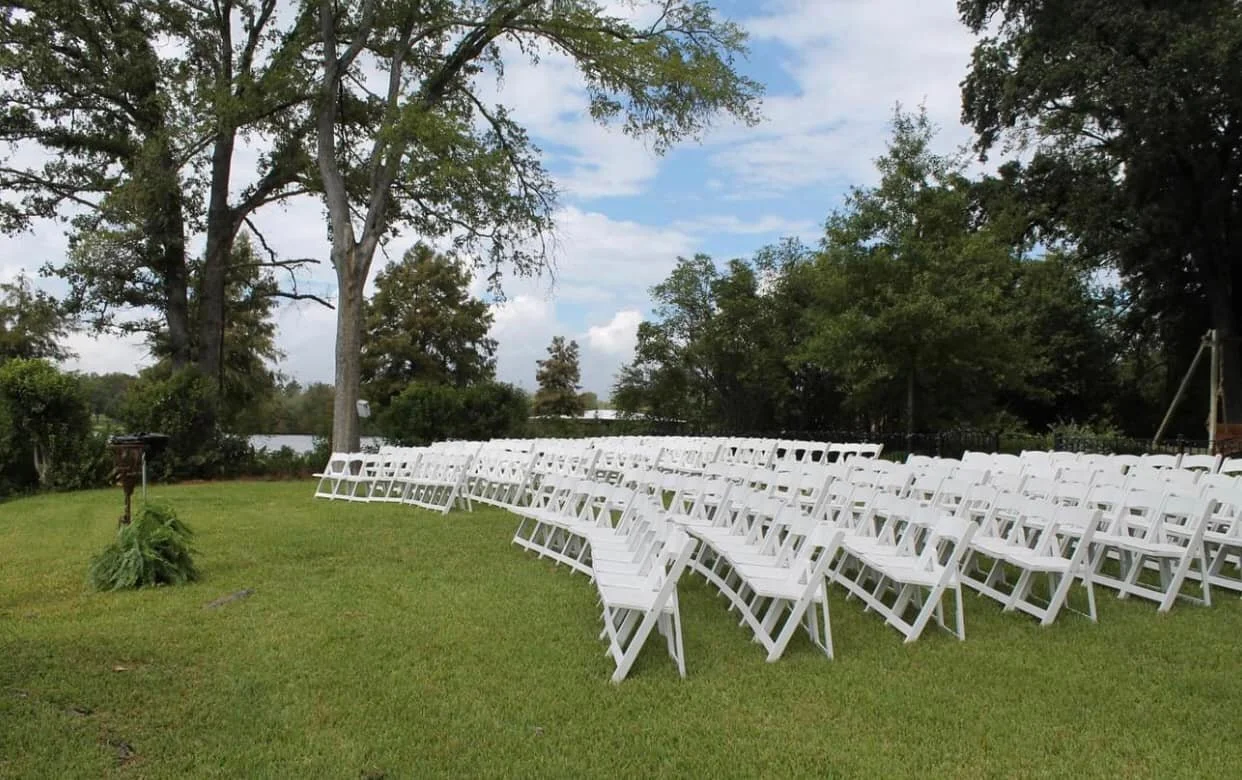 Rows of white folding chairs arranged outdoors on a grassy lawn, with trees in the background, suggesting a wedding or outdoor event setup.